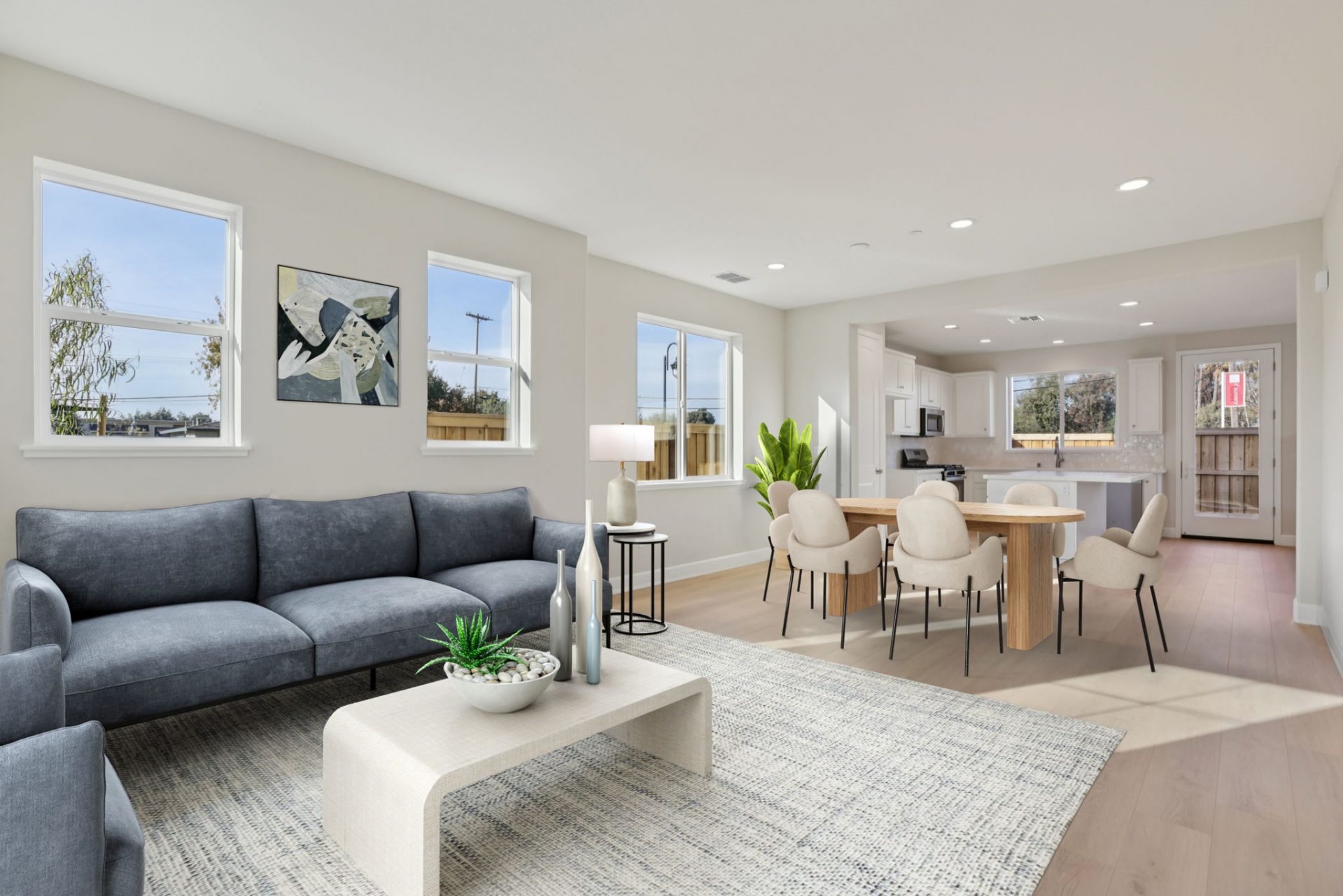 A virtually staged open concept room with a seating area furnished with a blue couch, coffee table and area rug. A dining table with six chairs complete with a plant in the corner along with other décor items.