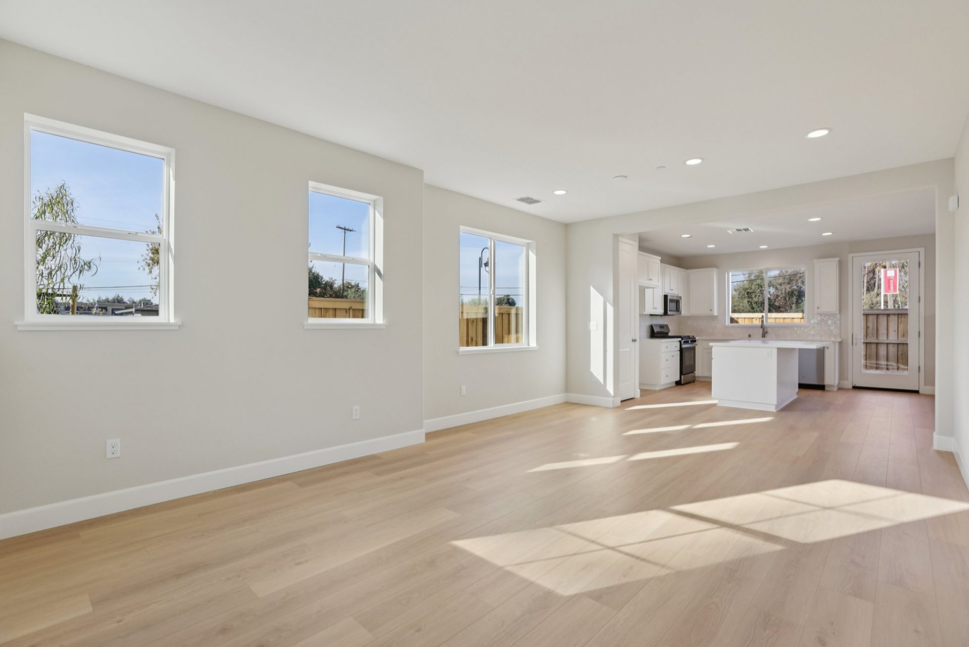 An unfurnished room with multiple windows, light wood flooring, an open concept space with a kitchen seen in the distance.