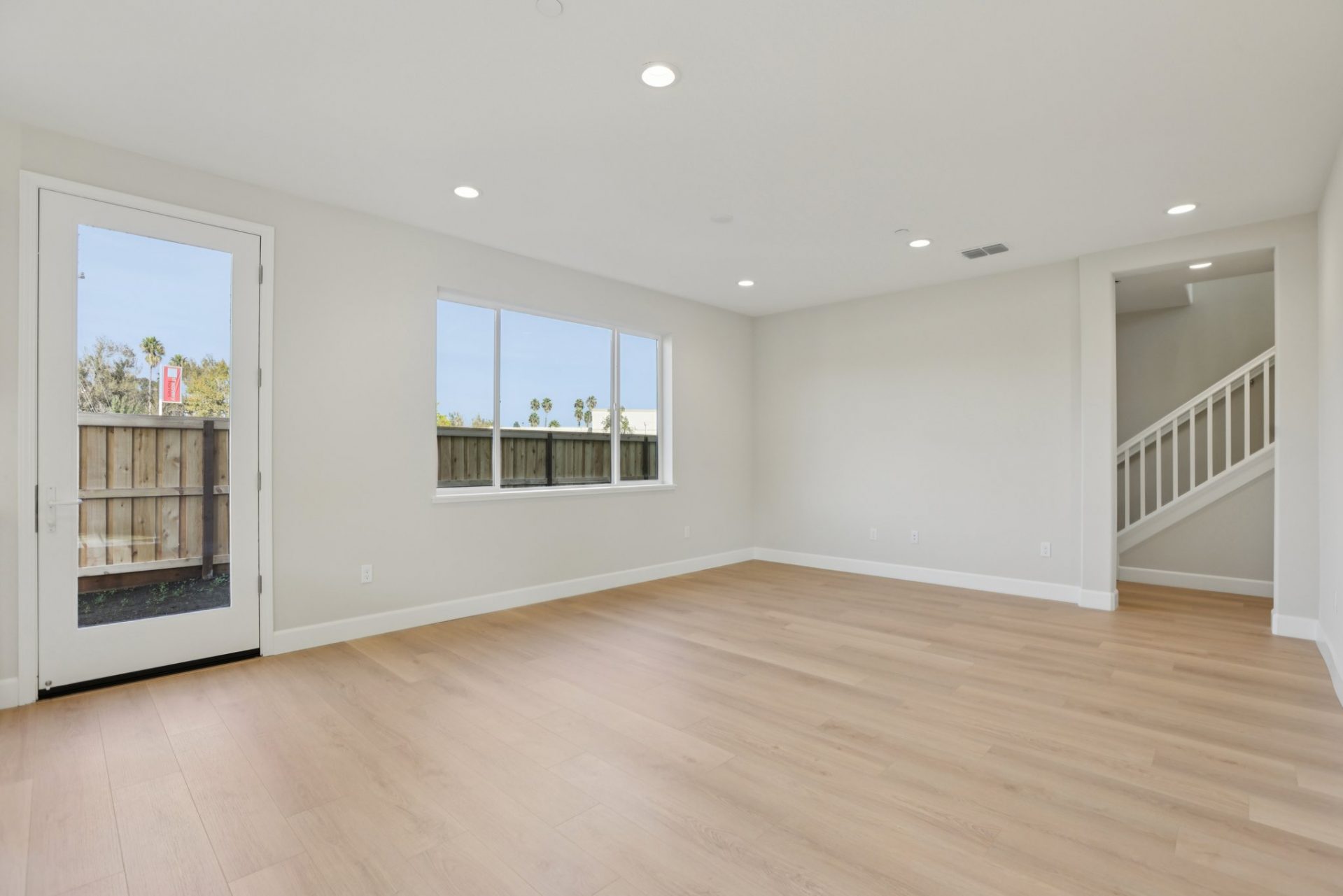 An unfurnished room with light wood color flooring, medium beige/gray walls with a glass panel door, a window, and can lights in the ceiling. To the right a staircase in the distance.