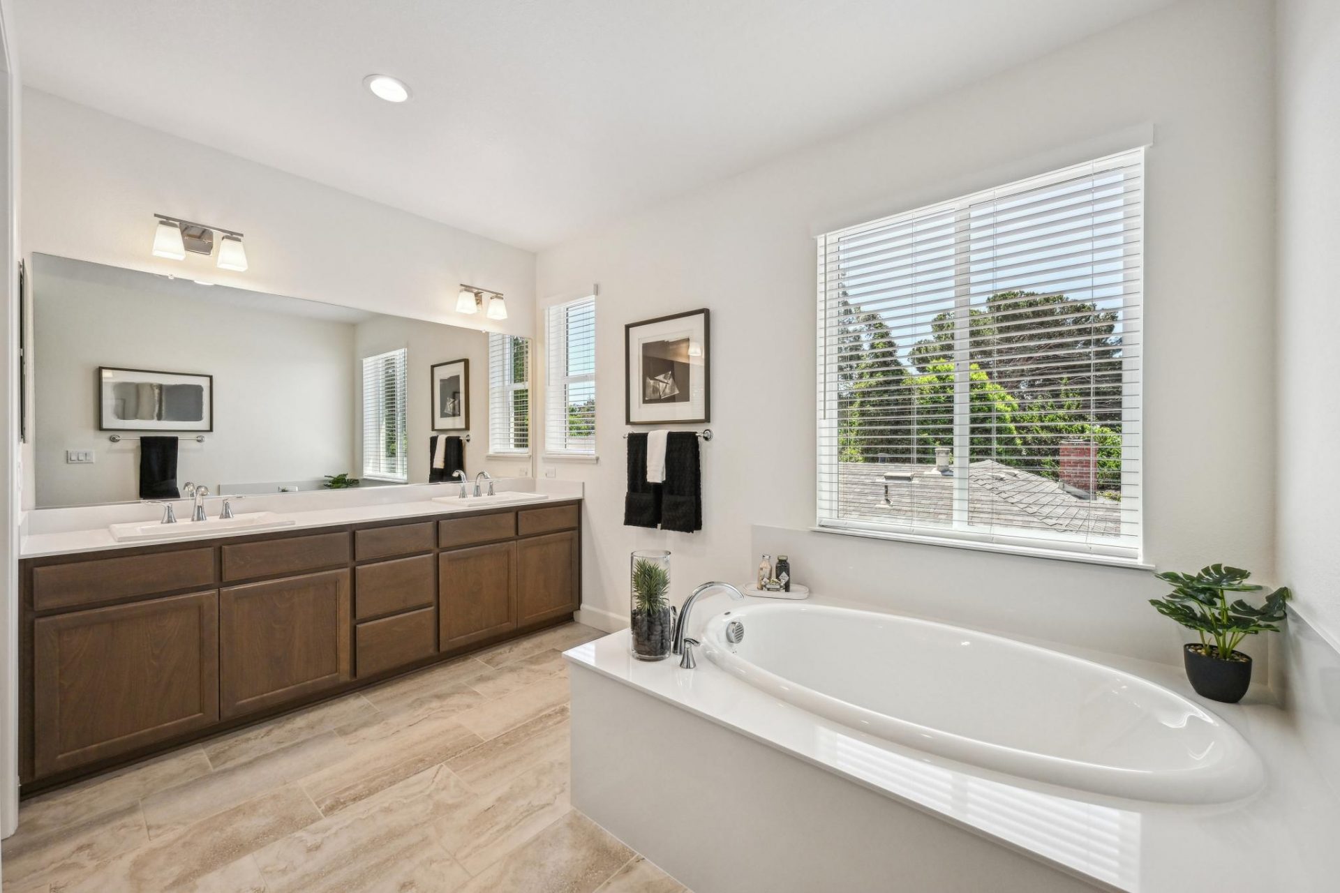 A primary bathroom with light colored tile flooring a white soaking tub with chrome faucet located underneath a window with a long double sink vanity, with chrome faucets, brown stained cabinets and a long mirror spanning the length of the wall. There are