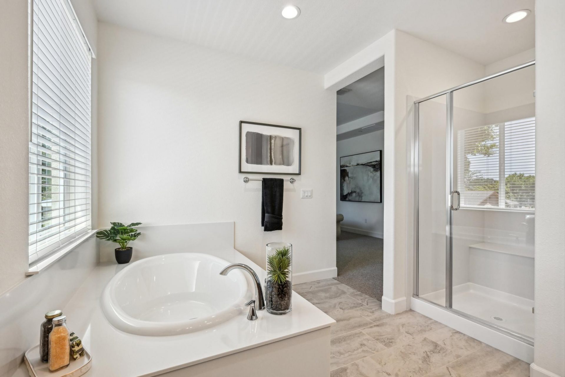 A primary bathroom with light colored tile flooring a white soaking tub with chrome faucet located underneath a window, and a glass enclosed shower.