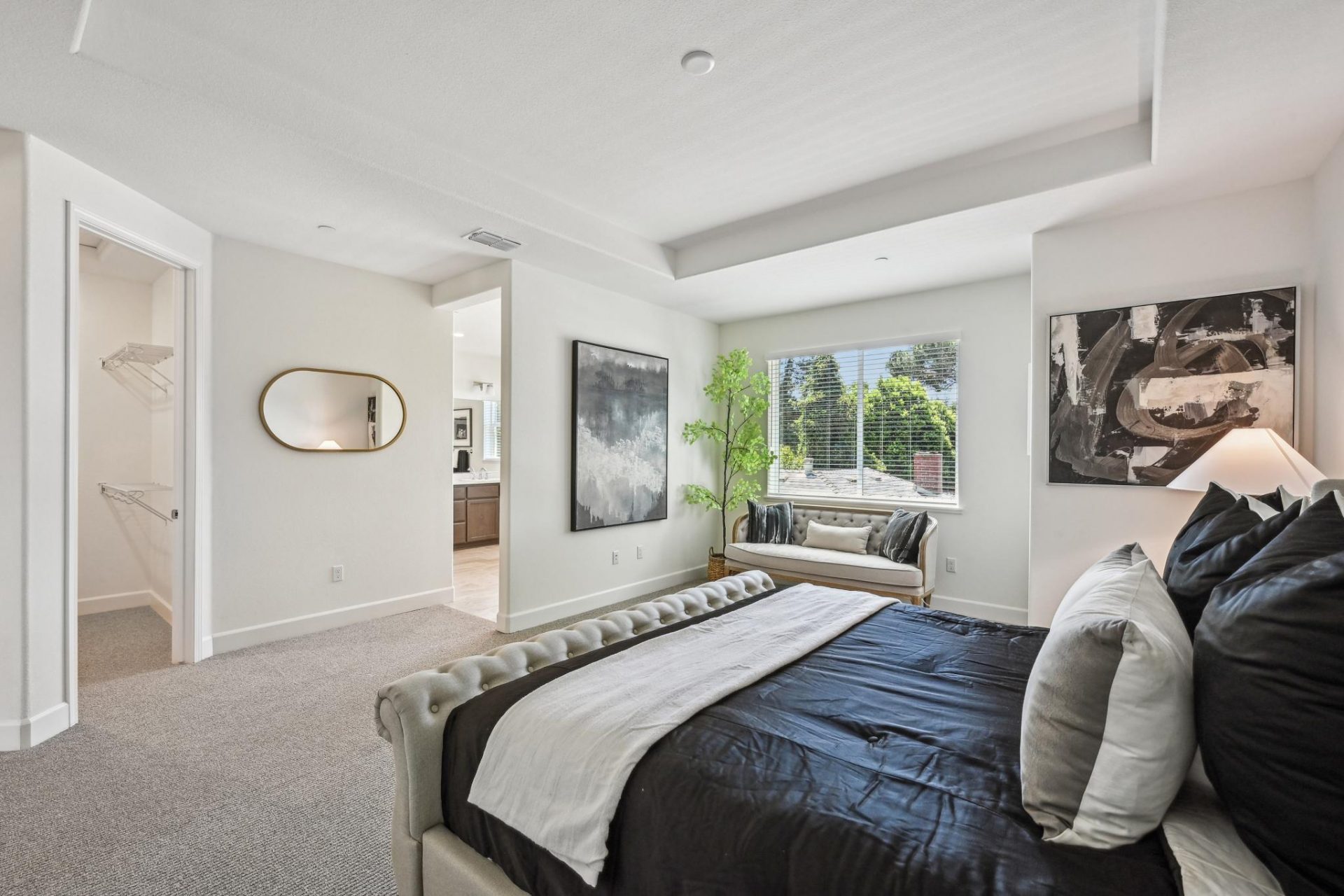 A large primary bedroom staged with a large king bed on a headboard and rails. To the right a seating couch under a window, with a large potted plant. The primary bathroom can be seen through an open entry way and a large closet is to the left.