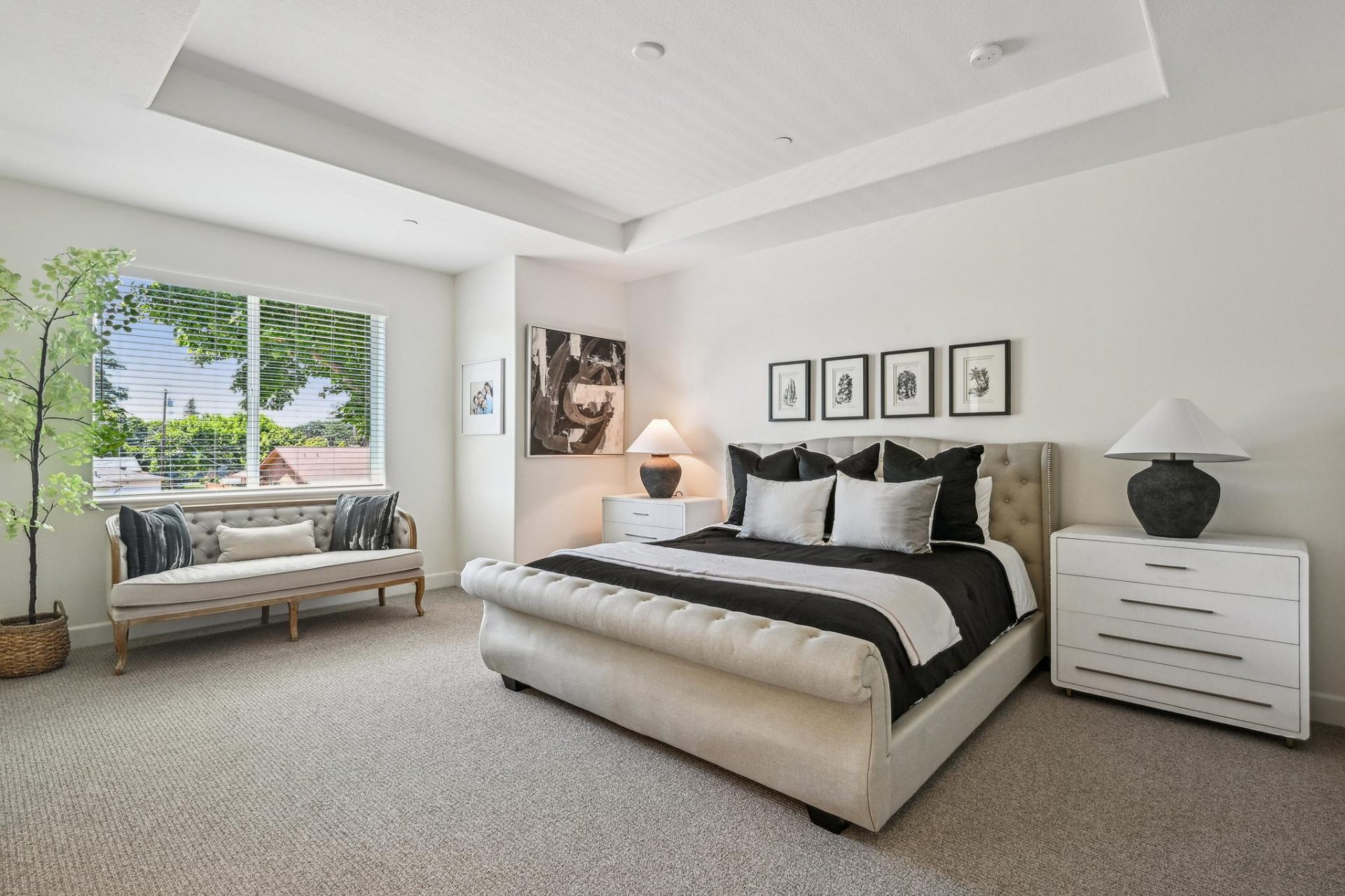 A large primary bedroom staged with a large king bed on a headboard and rails. To the left a seating couch under a window, with a large potted plant. Next to the bed are two nightstands with lamps and wall art. The carpet is medium gray and the walls are 