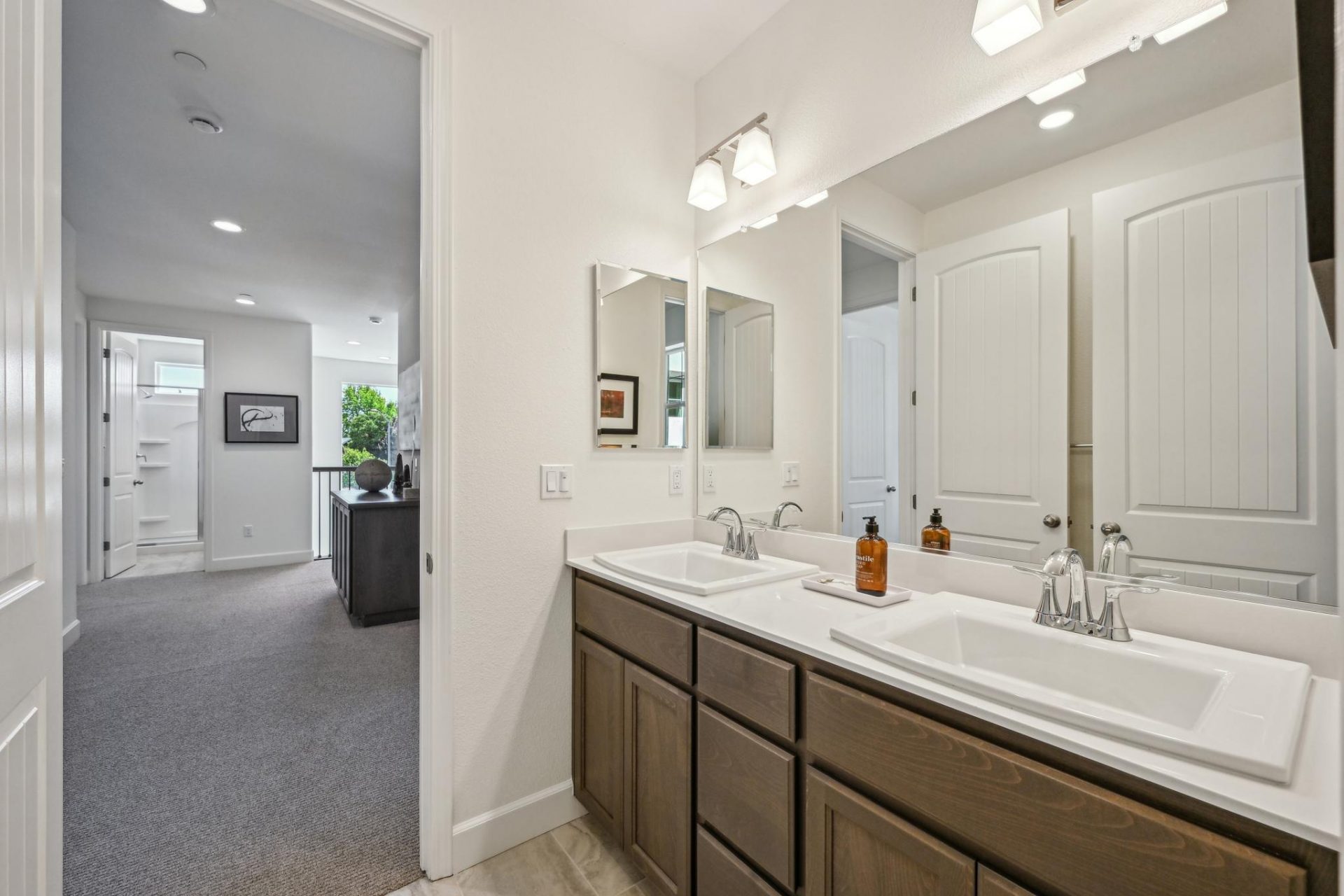 A bathroom next to a view of the stair landing space to the left. In the bathroom a two sink vanity with brown stained cabinets, and chrome faucets. A large mirror spans the space of the wall and two light fixtures located above.