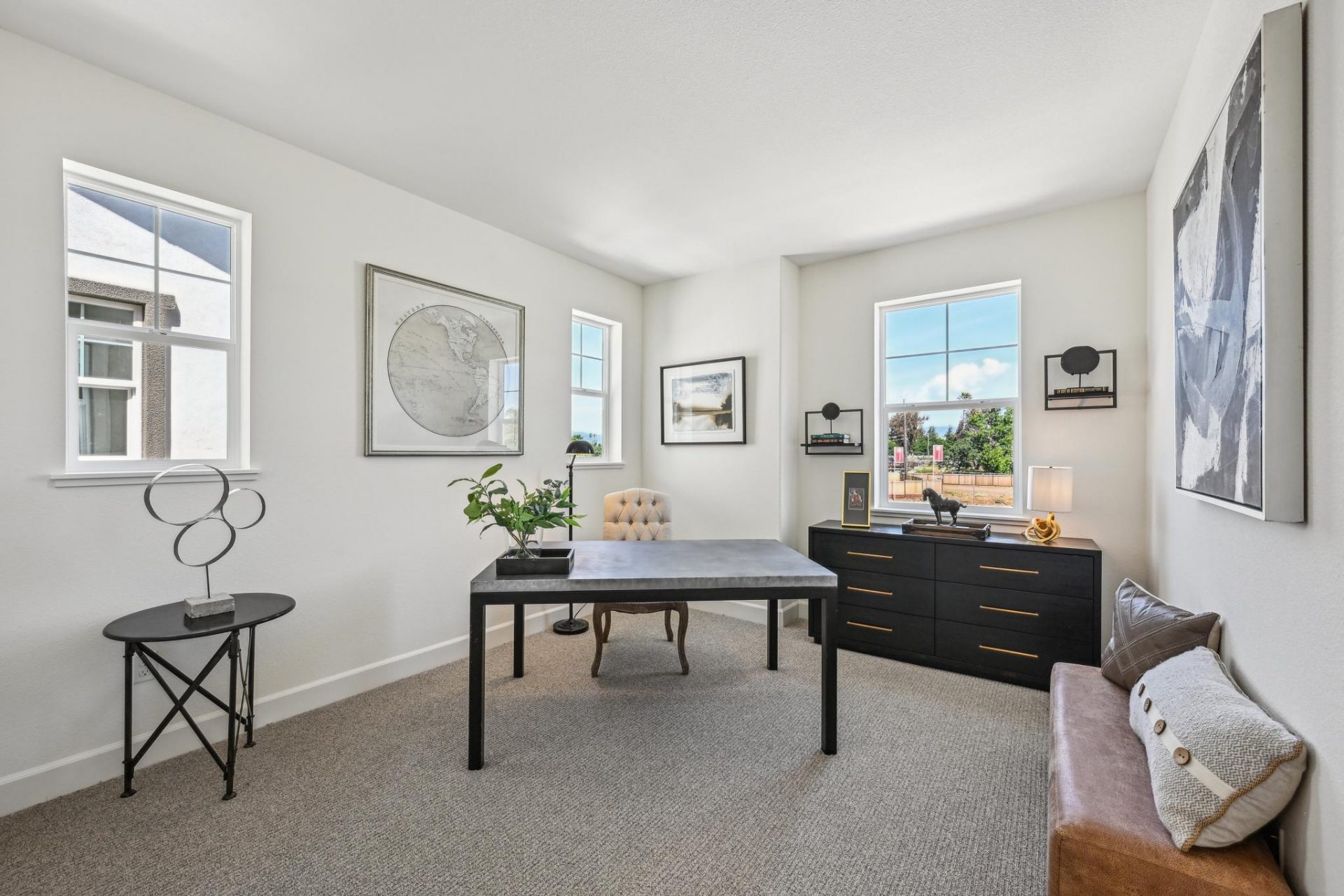 A room staged as an office with a desk and dresser , lighting, wall art and other décor items. Gray carpet and light colored walls with three windows brining in abundant lighting.