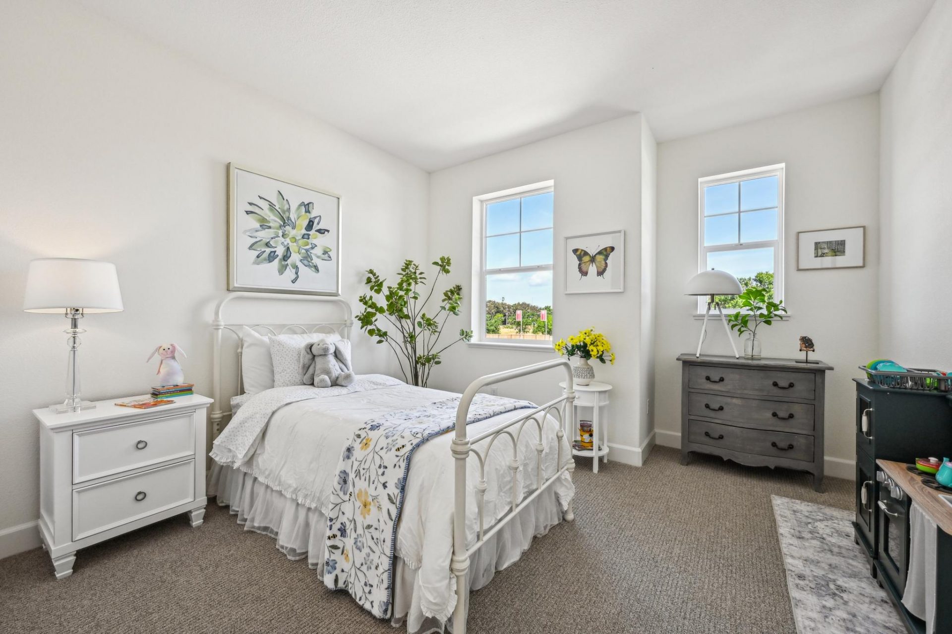 A bedroom staged for a young girls room with white and gray bed and dressers with white linens and flowers and butterfly accents. Gray carpet. light color walls and two windows provide a bright open space.