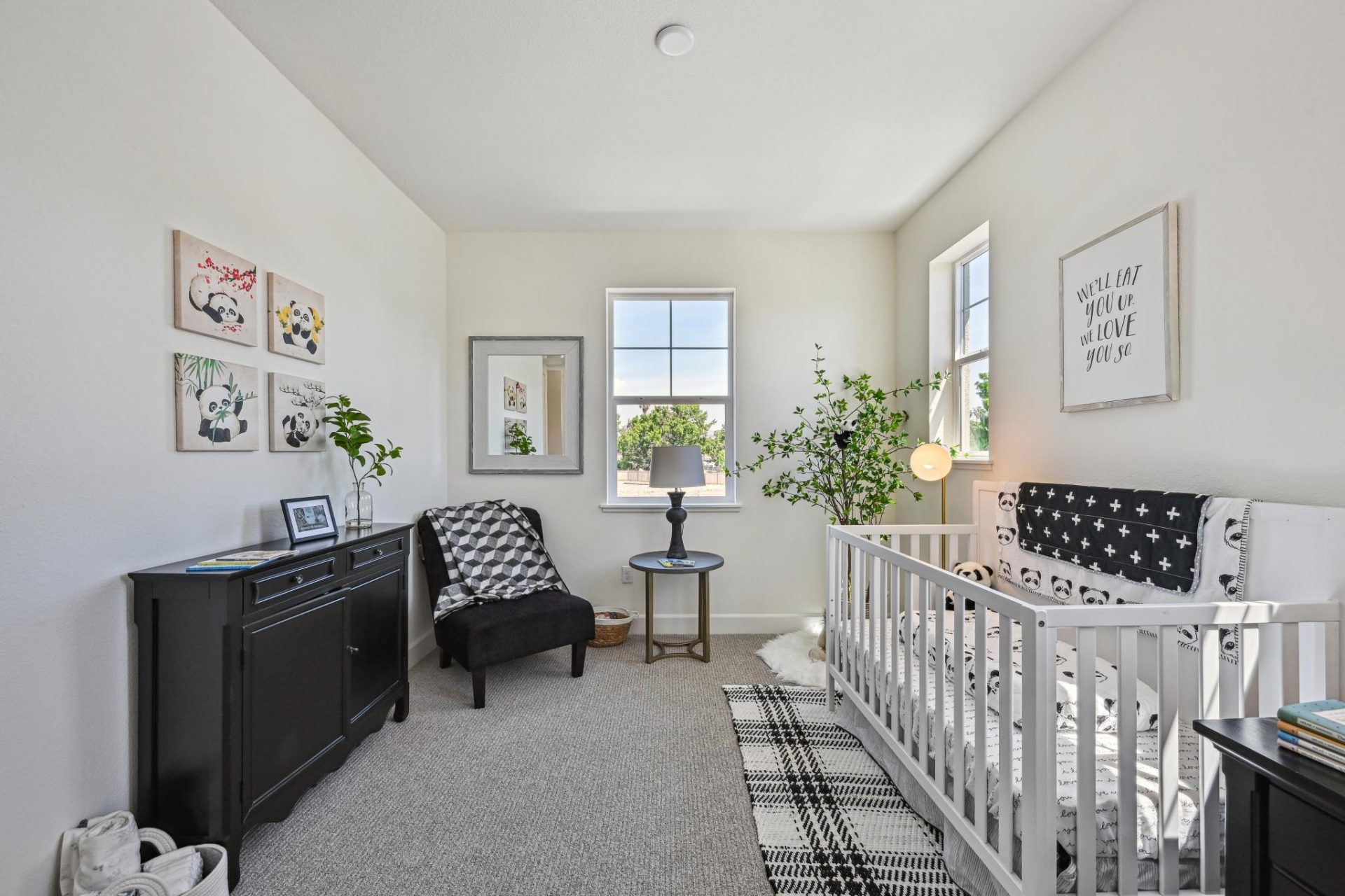A bedroom staged for a nursery space with light colored walls, gray carpet and two windows.  Black and white nursery décor with a panda theme and a potted green tree in the corner with other décor items throughout the room.