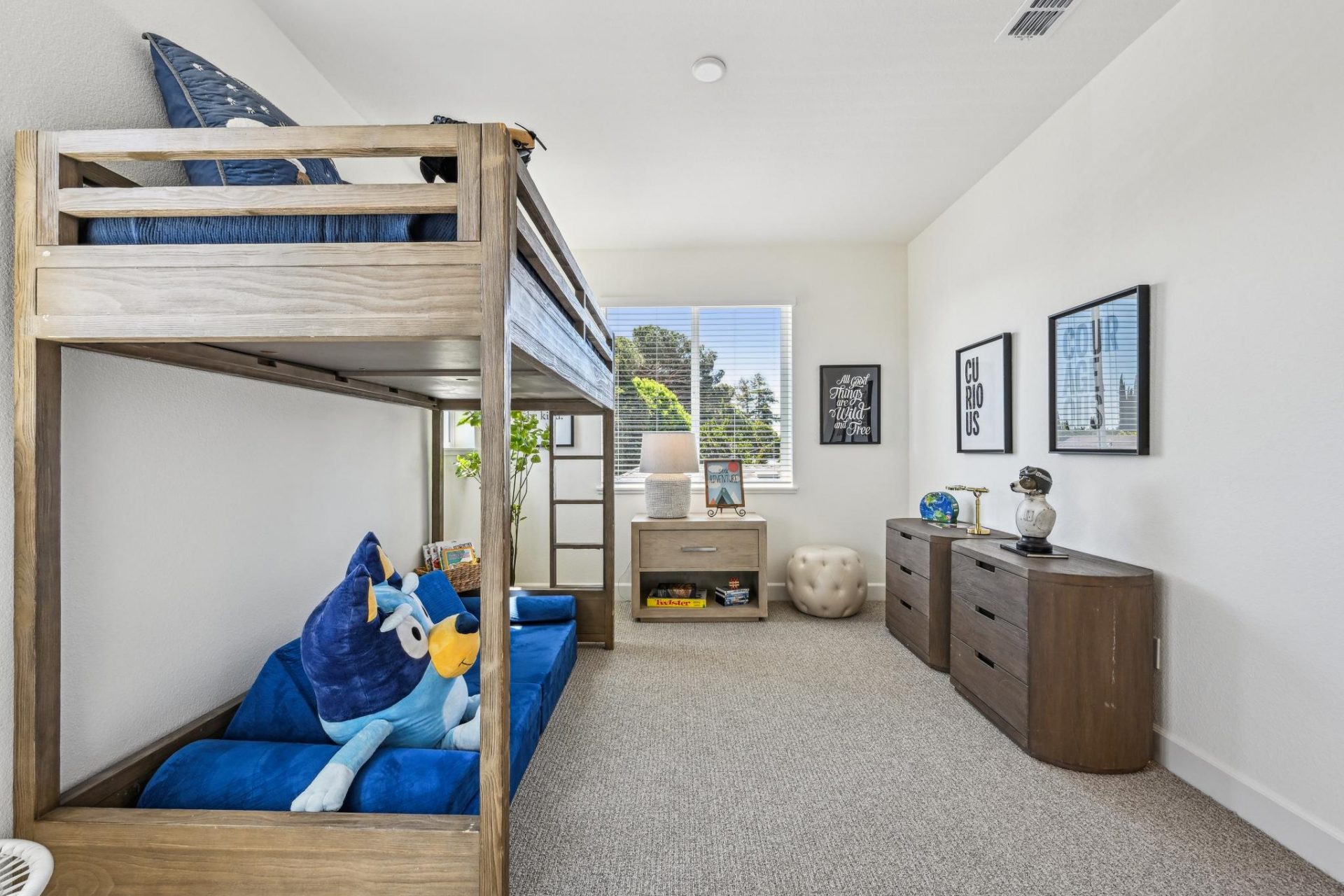 A bedroom furnished as a kids room with gray carpet, light color walls, and brown bedroom furniture including a bunk bed and dressers. There is a large window providing nice light into the space.