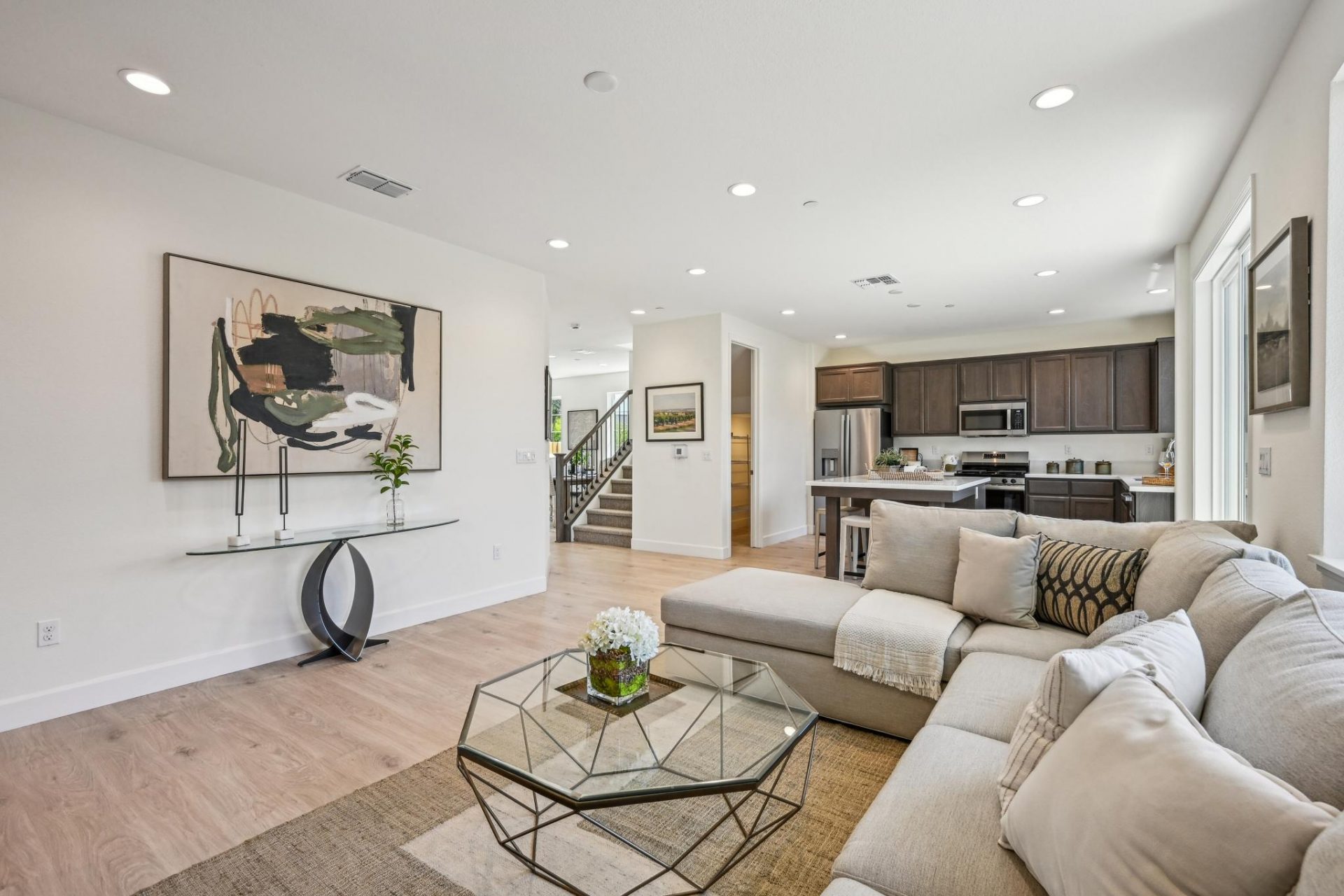 A furnished seating area with a large sectional couch and throw pillows and blankets, along with a glass coffee table and area rug, The floor is wooden and of light color, the space is open concept with the kitchen located across from this staged living a