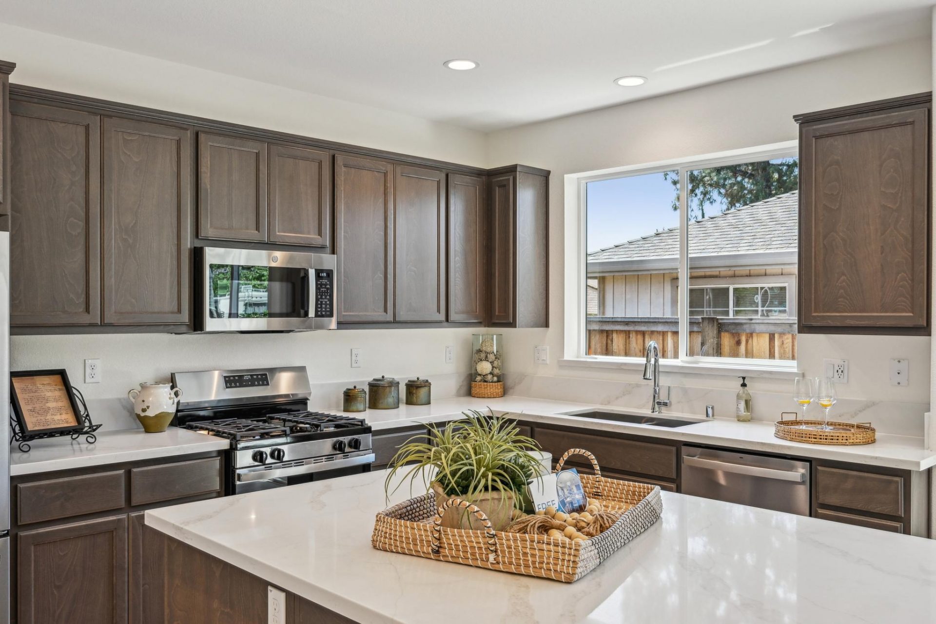 A modern kitchen with a stainless steel appliances, a gas range stove and microwave hood, brown stain color cabinets with light color countertops and a 6" backsplash. There is a dual space stainless steel sink with a chrome faucet with a window above it, 