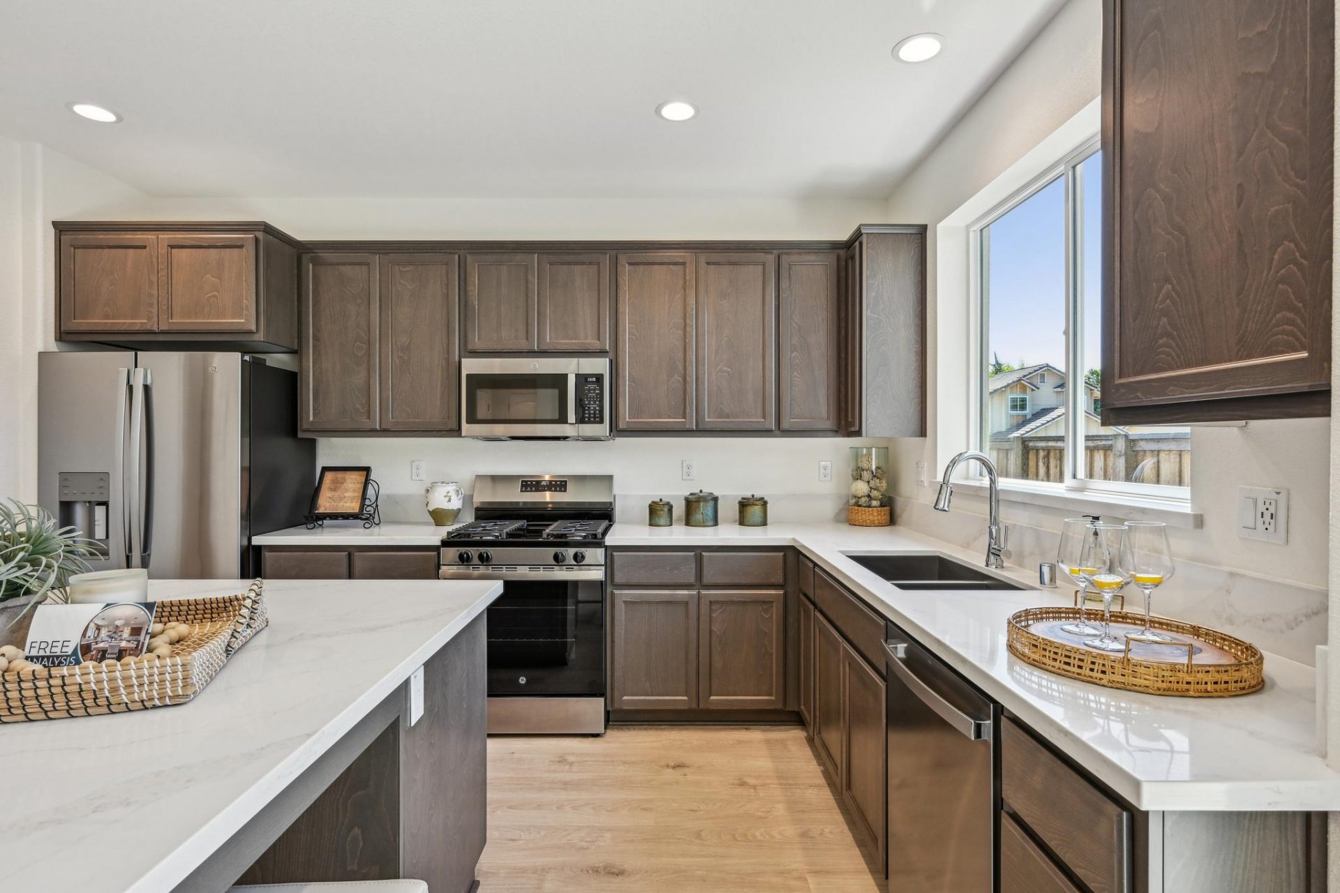 A modern kitchen with a stainless steel appliances, a gas range stove and microwave hood, brown stain color cabinets with light color countertops and a 6" backsplash. There is a dual space stainless steel sink with a chrome faucet and large kitchen island