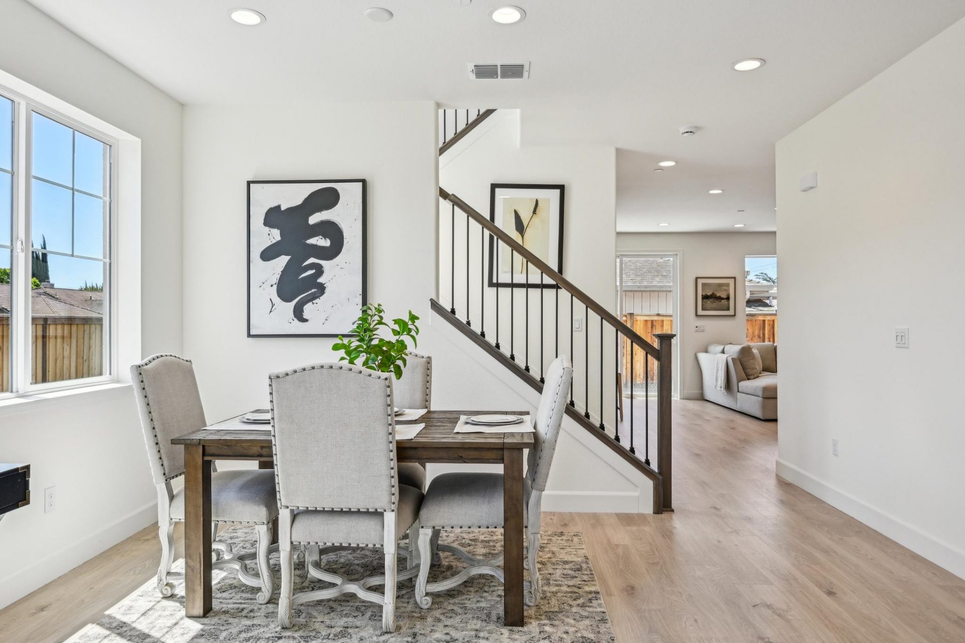 A staged dining room area next to a stair case, with a dining table and four chairs, light walls and light brown floors, with recessed can LED lights in the ceiling.