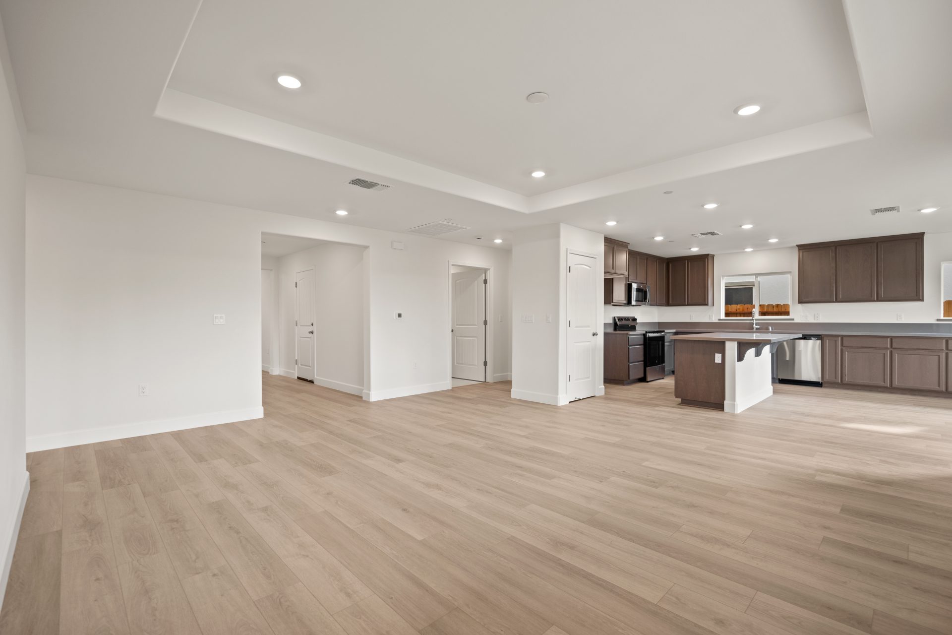 An unfurnished open concept area with light wood flooring, light walls and can lighting in the ceiling. A kitchen can be seen off to the right.