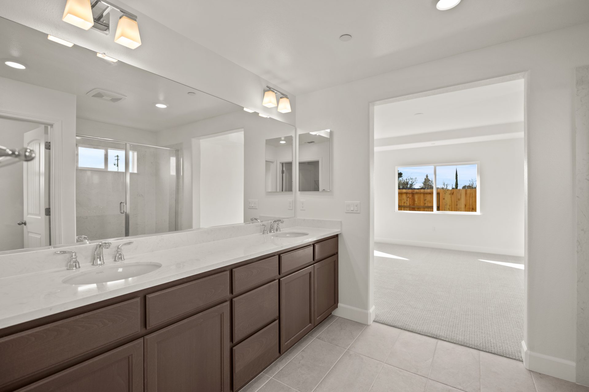 A two sink bathroom vanity with brown stained cabinets, and a light counter top. Light color tile flooring, chrome sink faucets, a long mirror and two light fixtures. View of the primary bedroom in the background