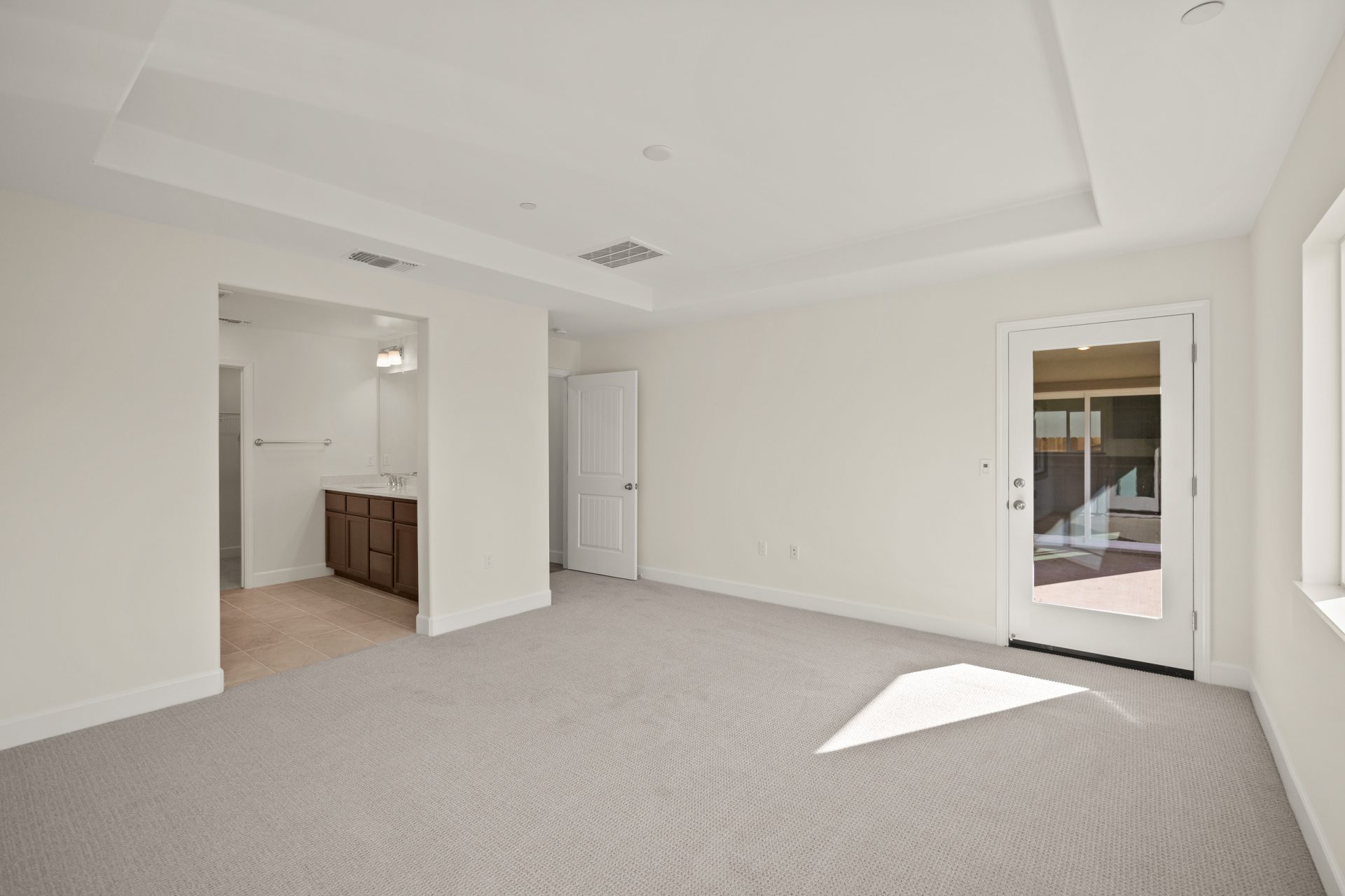 An unfurnished primary bedroom with gray carpet, light color walls, an inset high ceiling, and a windowed door leading to outdoor patio. To the left is the connected bathroom.