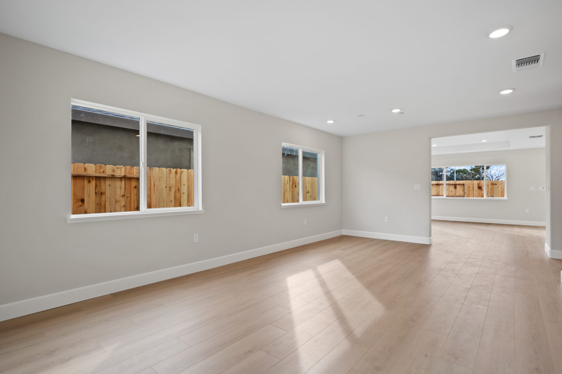 An unfurnished room with light brown wood color flooring, medium khaki color walls, 3 windows and a door, with white ceilings and recessed can lighting.