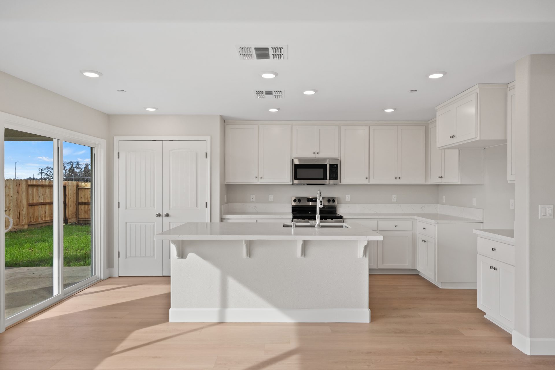 A modern kitchen with light color upper and lower cabinets, light colored countertops with stainless steel appliances, chrome sink faucet and a double door pantry.
