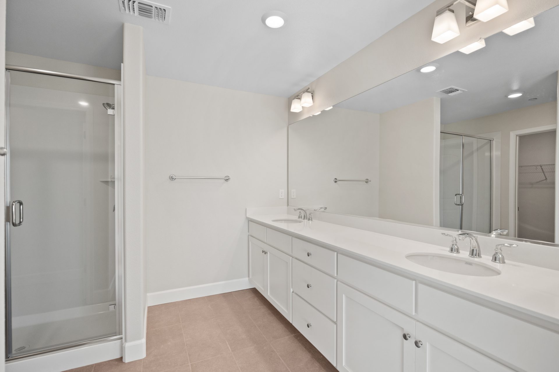 A bathroom with brown tile flooring, a dual sink vanity with white cabinets with nickel pull knobs, white countertops, chrome sink fixtures and a large mirror. To the left a shower with glass door.