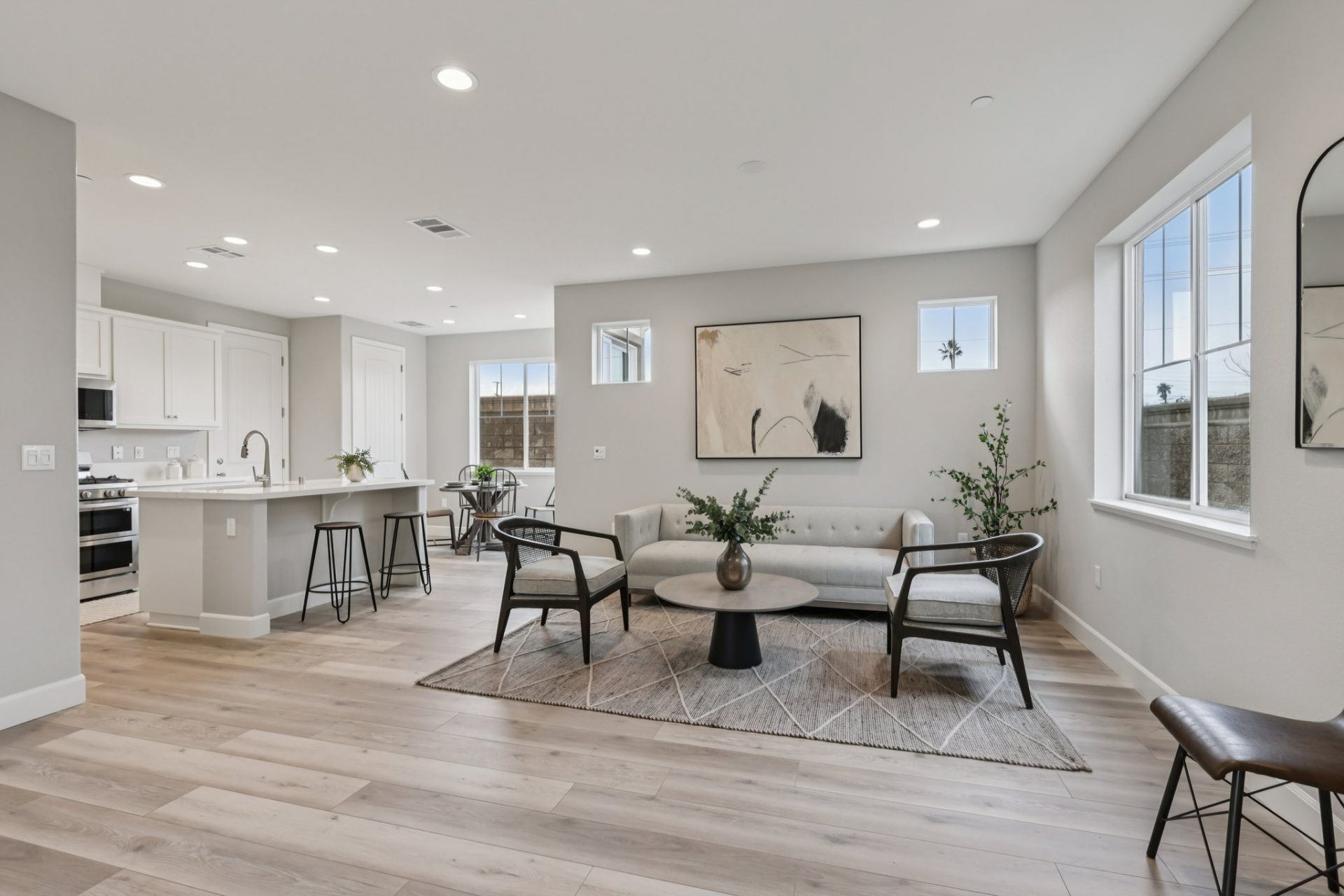 Interior open concept space with a living area furnished with a couch, chairs and coffee table. The kitchen is to the left , gray toned wood flooring, gray walls with white baseboards and LED can lights in the ceiling complete the space.