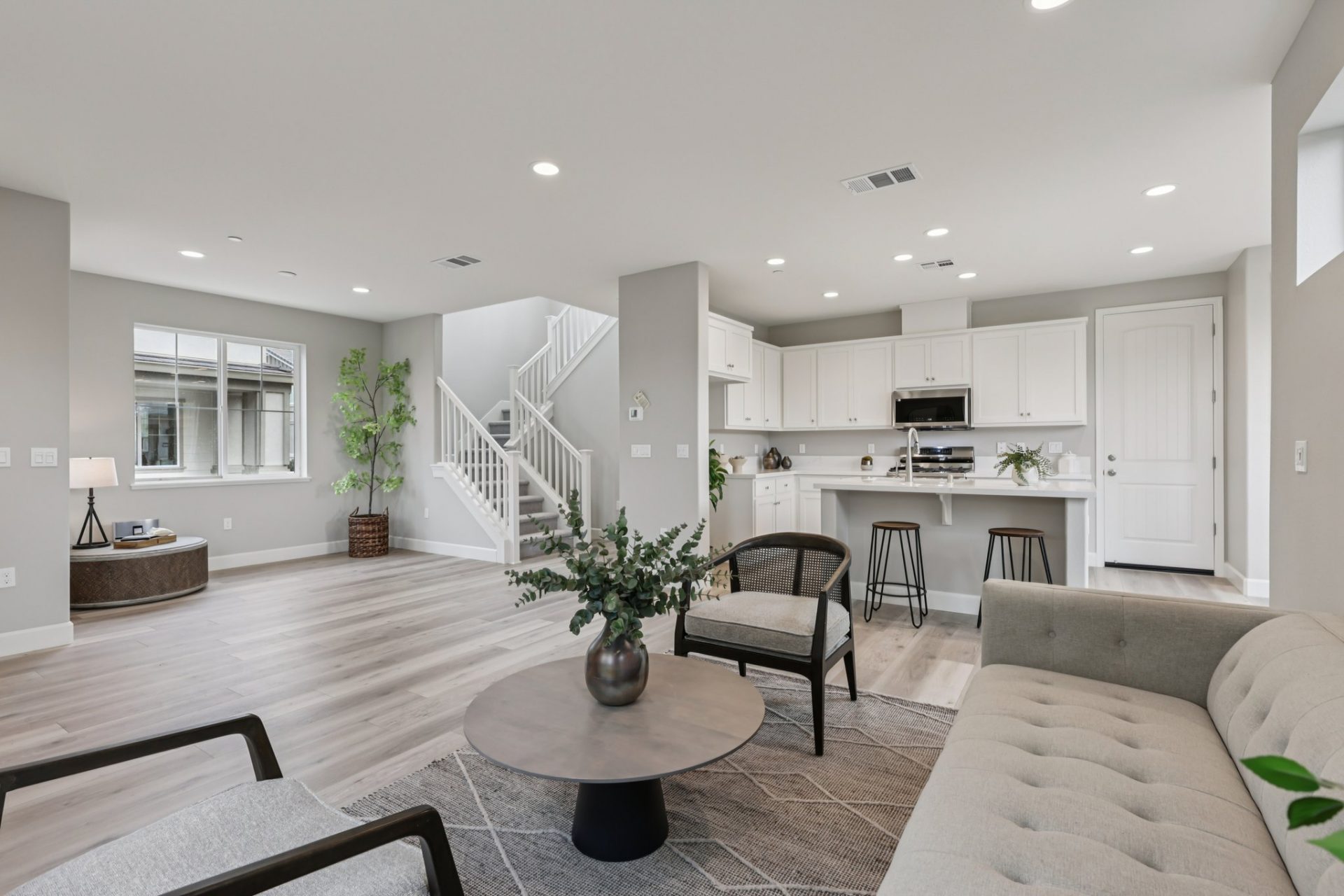 Interior open concept space with a living area furnished with a couch, chairs and coffee table. The kitchen is to the right and to the left a staircase along with decor and a window, light toned wood flooring, gray walls with white baseboards and LED can 
