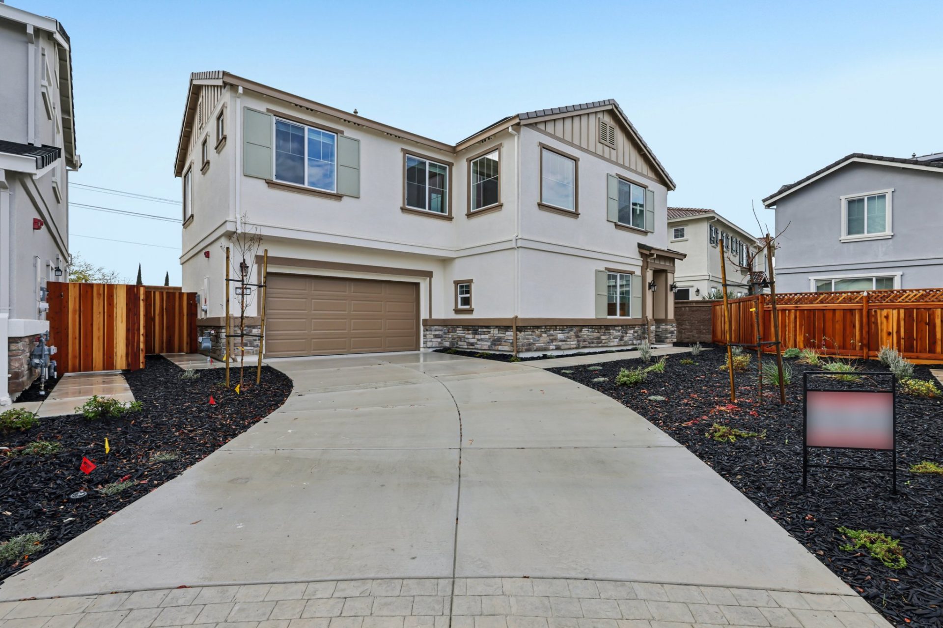 Exterior of a modern two story home with a tan and brown color scheme with brick detail on the bottom of the home, lots of windows and a two car garage. The yard is complete with dark mulch and shrubs.