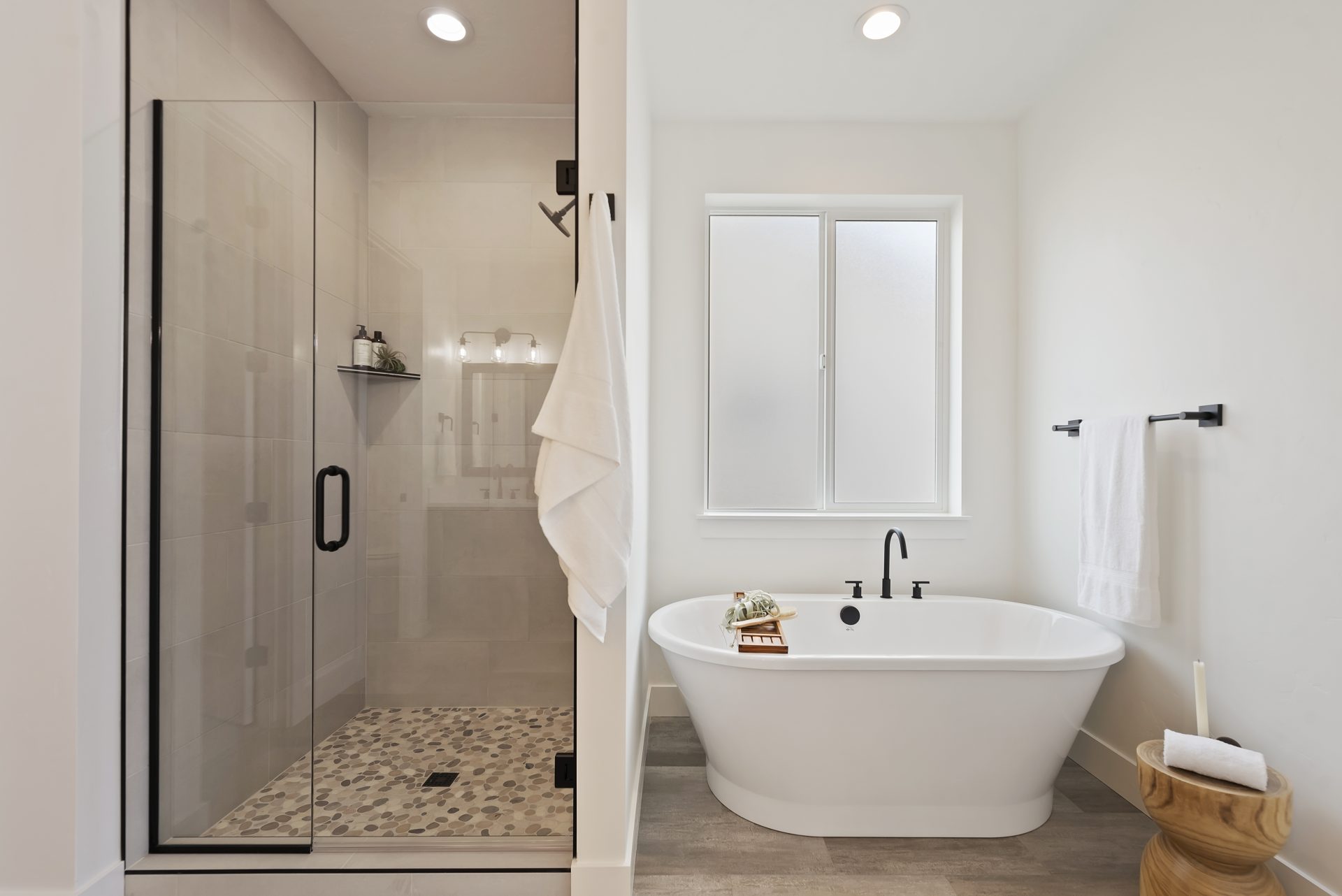 A bathroom with a glass enclosed shower on the left with black handles and finishes, a stone shower pan and tile from floor to ceiling. To the right of the shower, a white free standing soaking tub with black faucet and a frosted window located above.