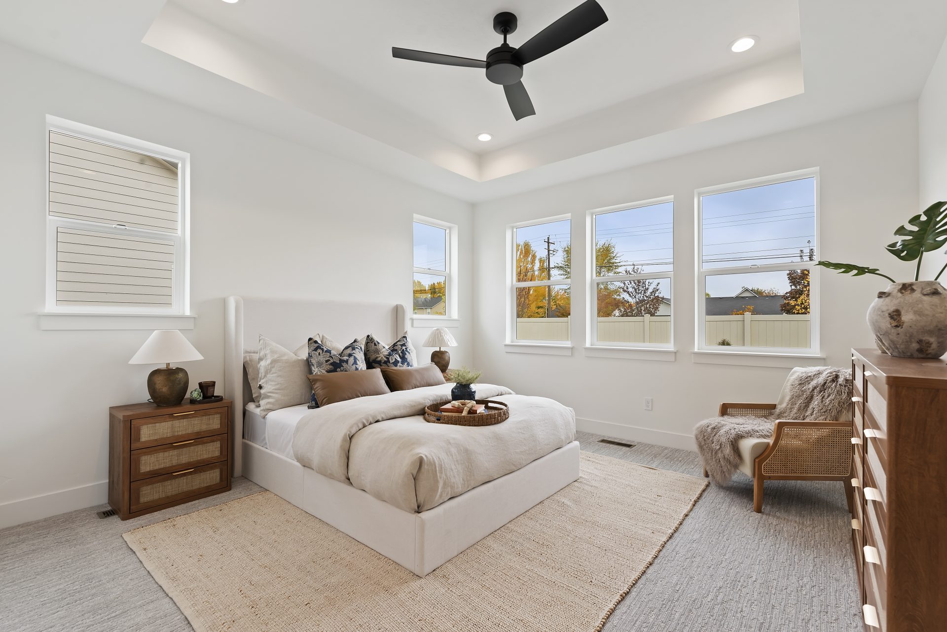 A staged bedroom with gray carpets, light color walls, 5 windows, and black ceiling fan and LED can lighting.