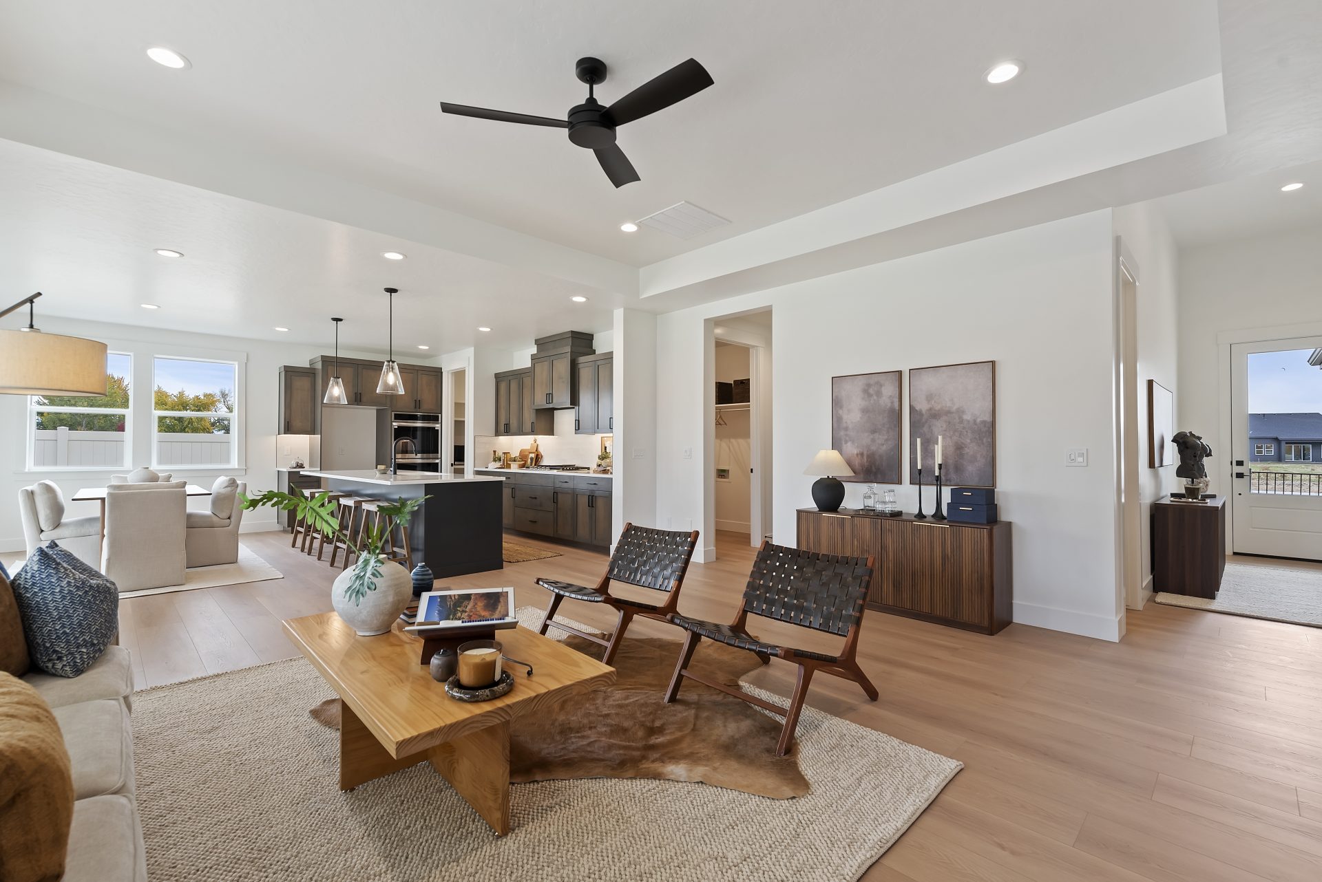 An open concept space with a  living room, dining room and kitchen, flooring is a light brown wood, the walls are a light color, and LED can lights in the ceiling. The living area is staged with a seating area, there is a ceiling fan, and other decor item
