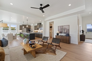 An open concept space with a  living room, dining room and kitchen, flooring is a light brown wood, the walls are a light color, and LED can lights in the ceiling. The living area is staged with a seating area, there is a ceiling fan, and other decor item
