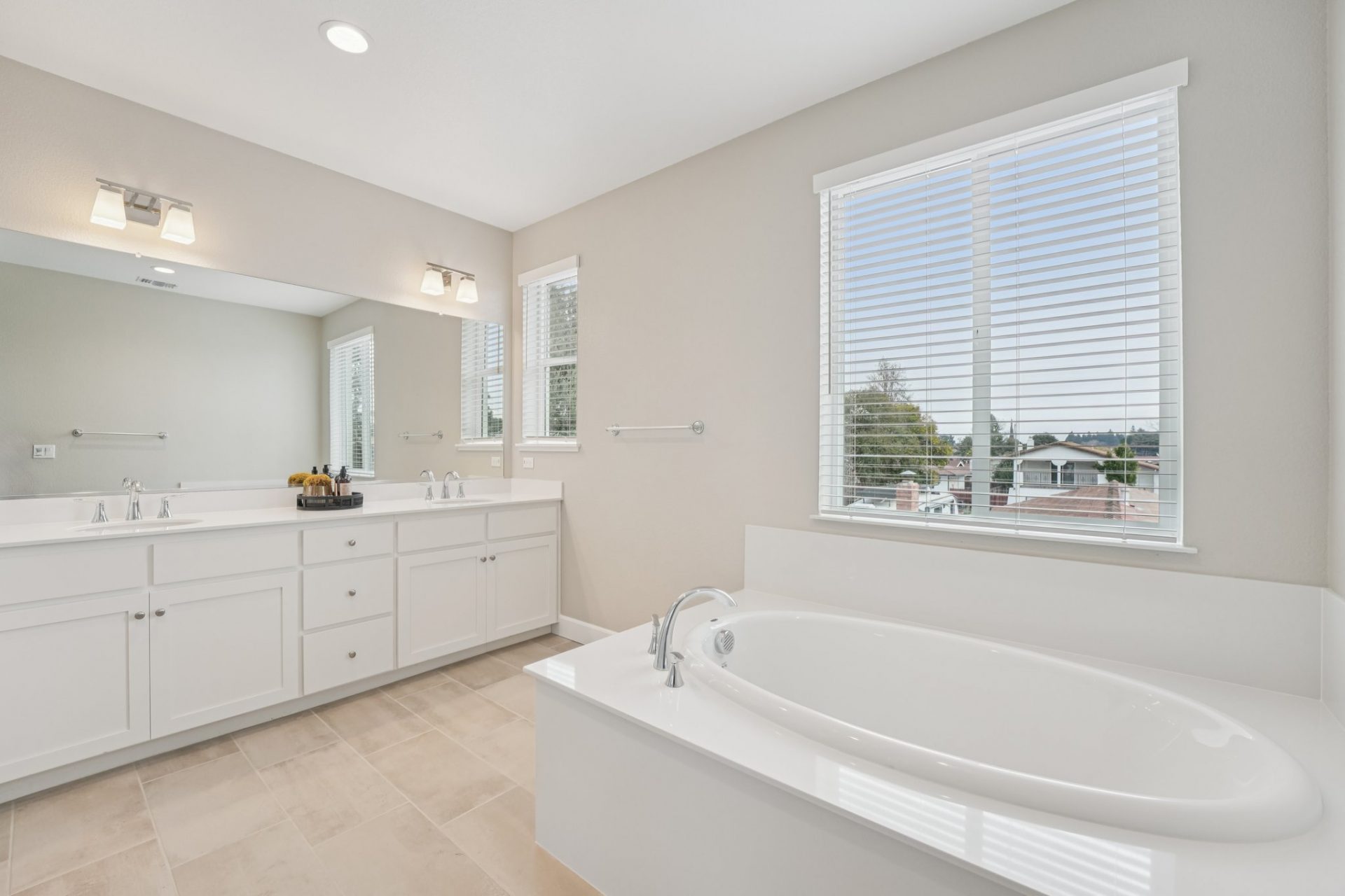 A bathroom space with beige floor tile, a white soaking tub to right, a double sink vanity with a mirror spanning the length of the vanity with two light fixtures above and two windows.