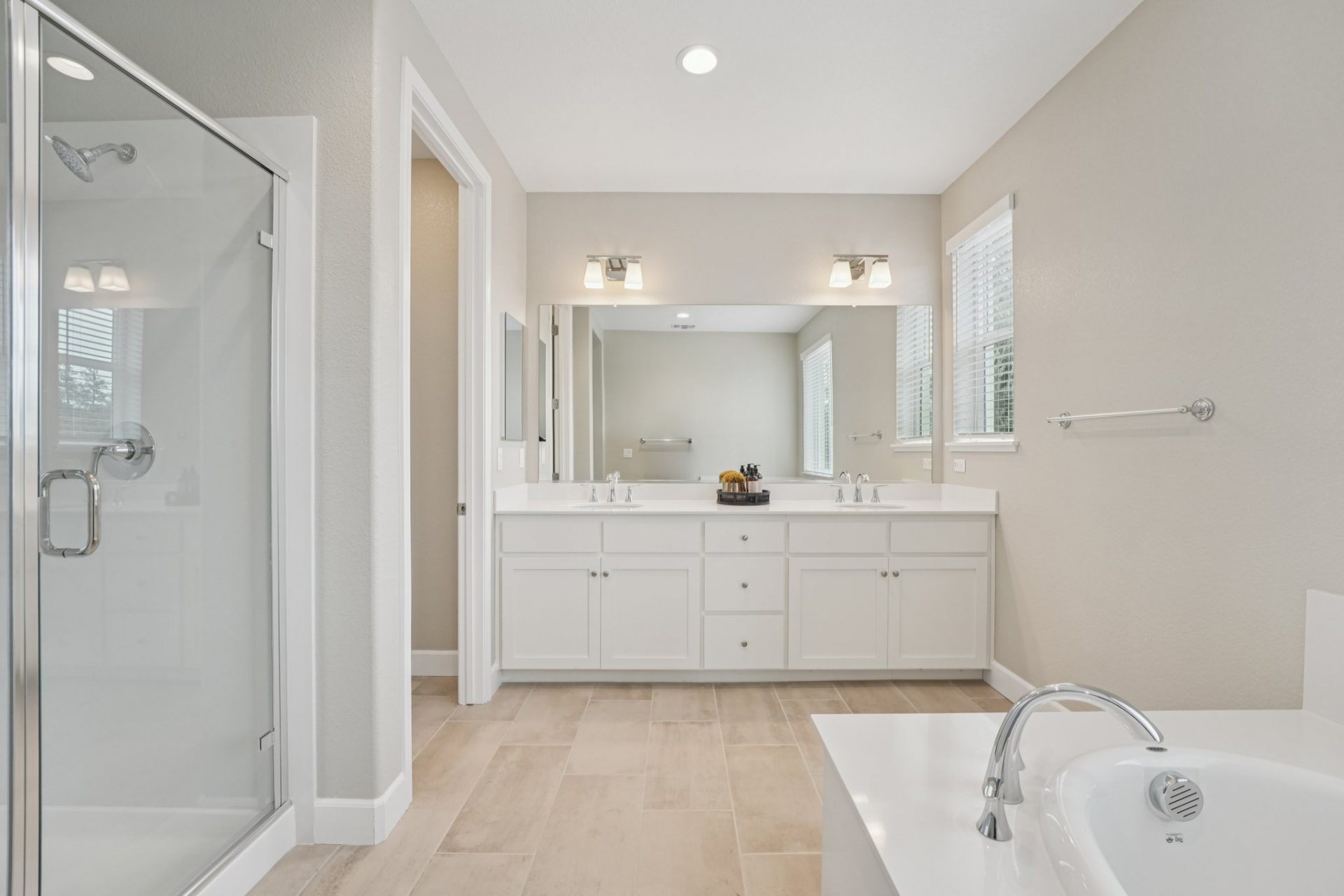 A bathroom space with beige floor tile, a white soaking tub to right, a glass enclosed shower to left, and straight ahead a double sink vanity with a mirror spanning the length of the vanity with two light fixtures above.