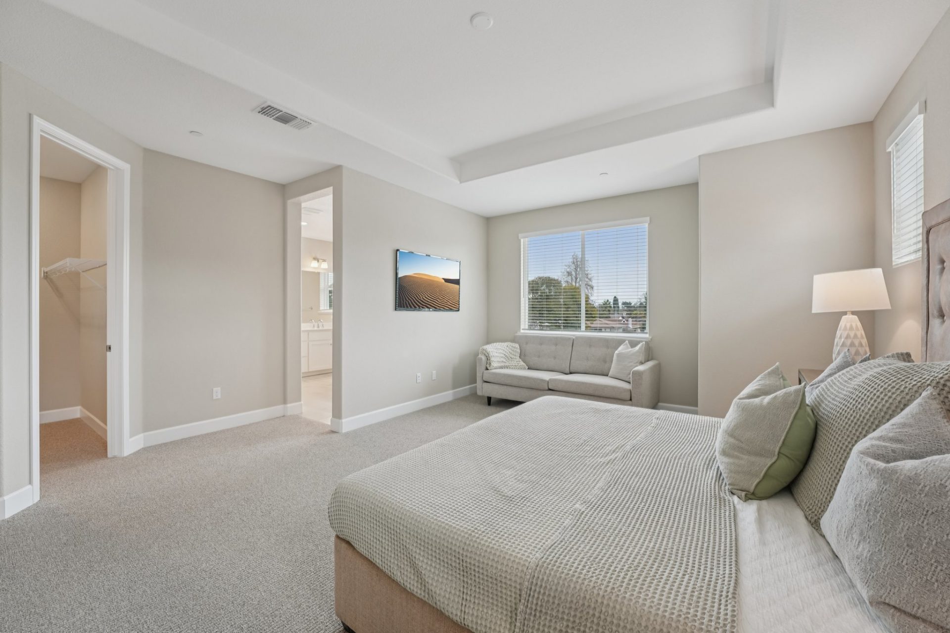 A large primary bedroom furnished with a bed set, a chaise under a window and nightstand with lamp. There is gray/ beige carpet with light beige walls, and a white ceiling with a fan prewire. Entry to the closet and bathroom can be seen.