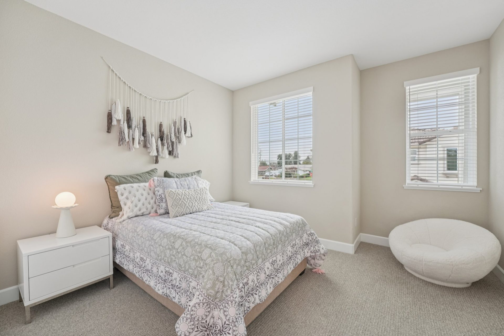 A furnished bedroom with light grey/beige carpeting, light beige walls, two windows and a white ceiling.  The room is furnished with a twin bed, two nightstands, a seating poof and wall decor.