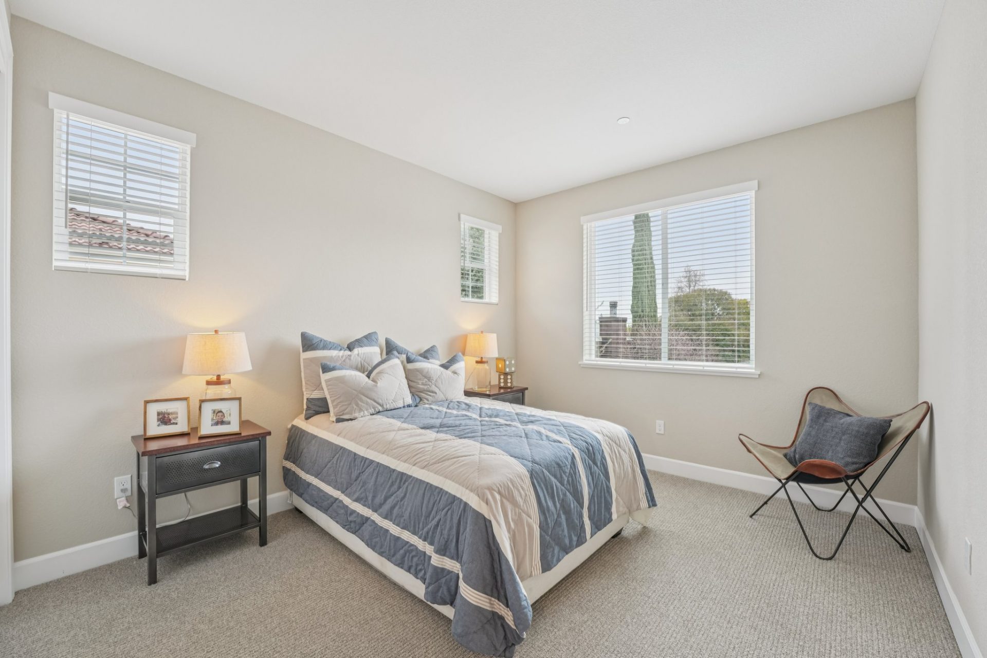 A furnished bedroom with light grey/beige carpeting, light beige walls, three windows and a white ceiling.  The room is furnished with a twin bed, two nightstands with decor and a side chair.