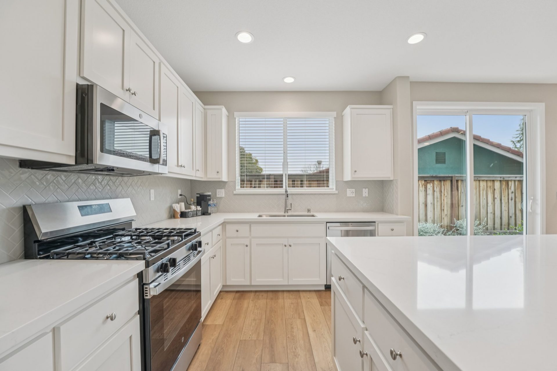 A kitchen space with white upper and lower cabinets spanning a fill wall, a sink and pull down faucet below a nice sized window. To the left are a gas range stove and microwave hood, and spanning the space of cabinetry is countertop to cabinet tile backsp