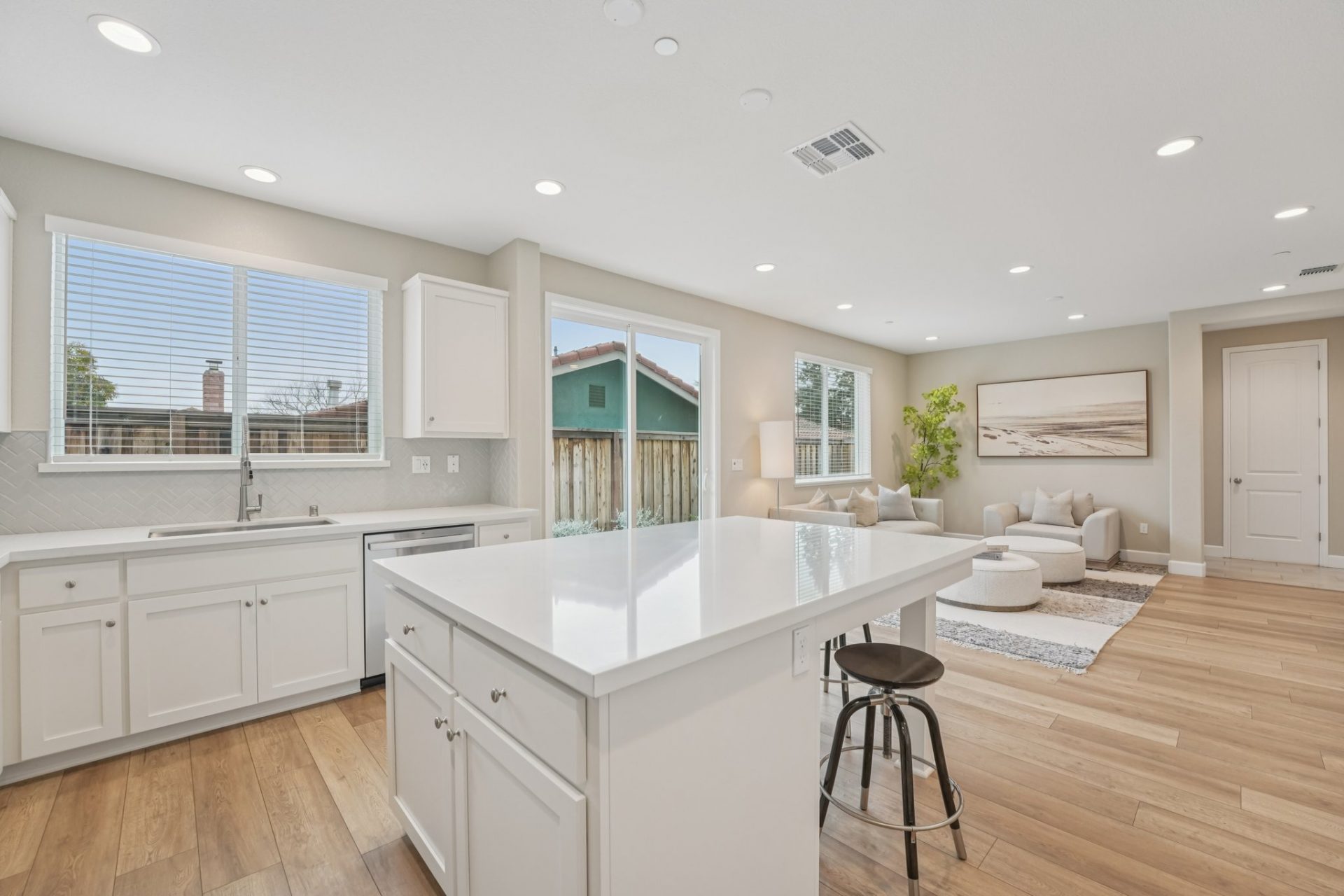 View over a kitchen island with white cabinets and nickel knobs, and a white countertop. Ahead is a furnished seating area with lots of windows and a sliding glass door leading to the back yard.