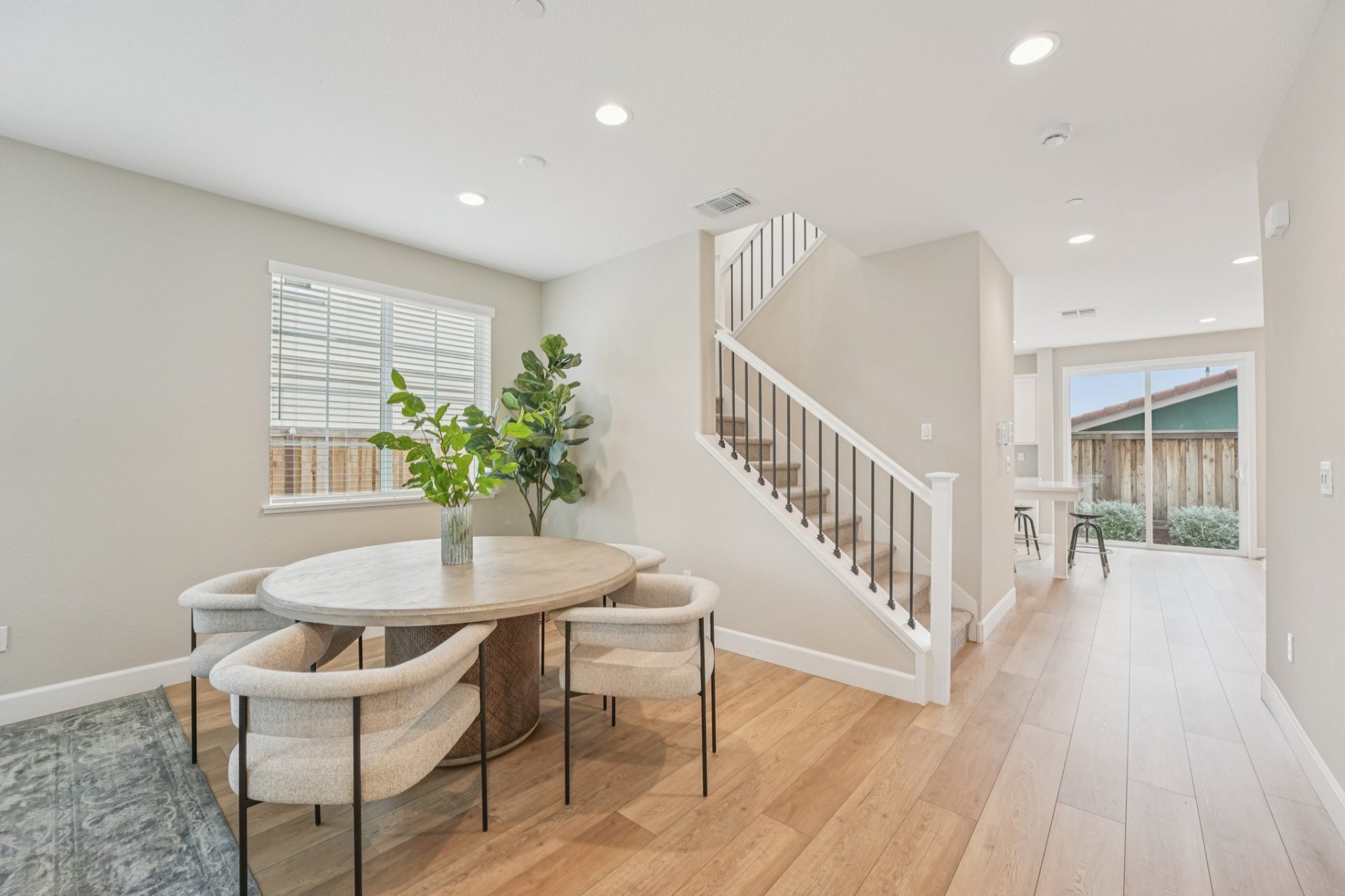 A dining area next to a carpeted staircase, with wrought iron balusters, and white handrails.  There is a round dining table with four chairs and green plant decor. The floors are a light wood, and the walls a light beige with a white ceiling and LED can 