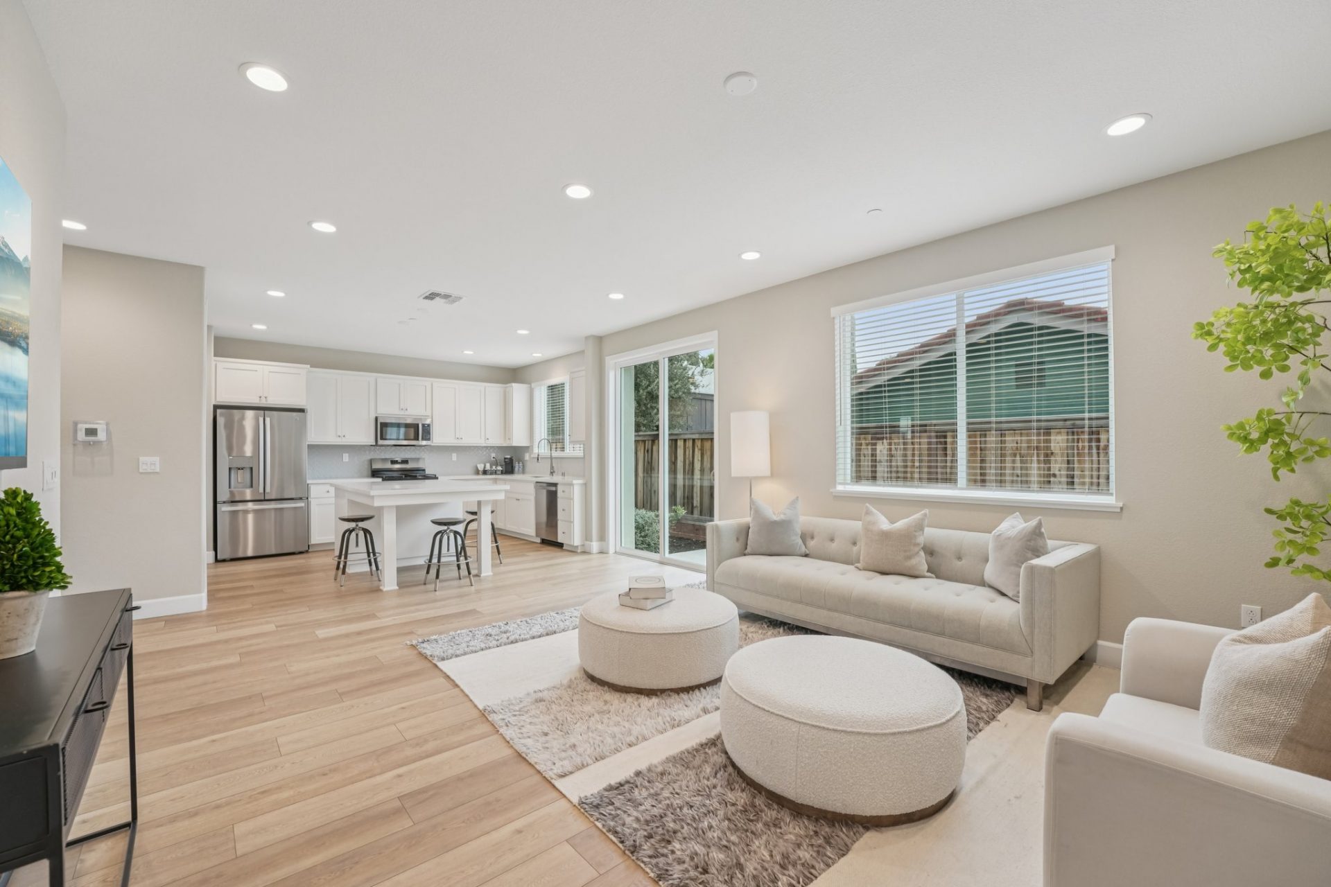 A furnished open concept living space with a kitchen in the background. Multiple windows and a slider provide ample light to the space, the floors are a light wood , light color walls and a white ceiling with LED canned lights.