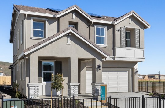 Exterior of a home with multiple roof peaks, with a 3 color paint scheme, with a mix of light and dark grays and an off-white color garage door. There are 3 pillars at the entry way with brick detail towards the bottom with mulch and plant landscaping.