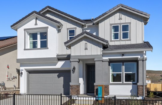 A front exterior of a home with stucco and wood paneling with a three color paint scheme from off- white to medium and dark gray with mulch landscaping with small plants. A tall covered entry way with pillars that have brown stone accents at the base.