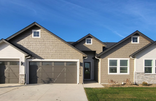 Exterior of a modern home with a tan, off- white and dark brown color scheme. Home has horizontal wood plank and wooden shingle architecture with 5 roof peaks, a 3 car garage, grass and tock landscaping.