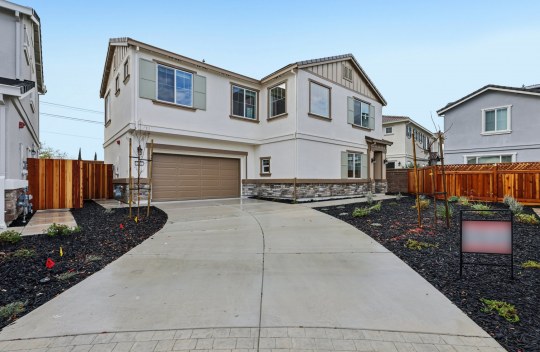 Exterior of a two story home with a multi color paint scheme including tan and brown with stone detail near the base of the home, and dark mulch landscaping with light shrubbery.