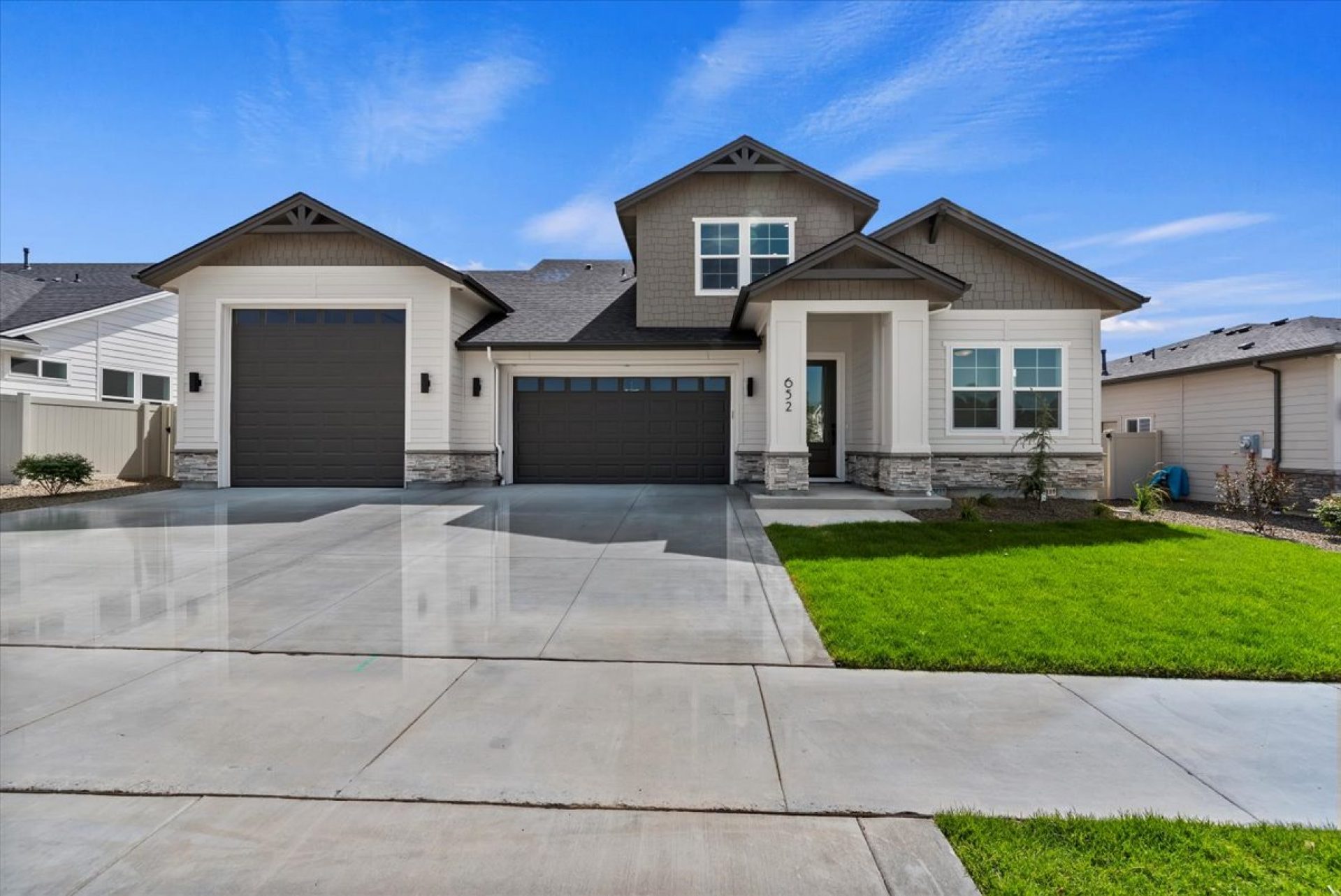 An exterior of a single story home with a gray and tan color scheme with dark accents of the roof and garage doors. There are stone details at the base of the home and green grass landscaping with rock pebbles and shrubs.