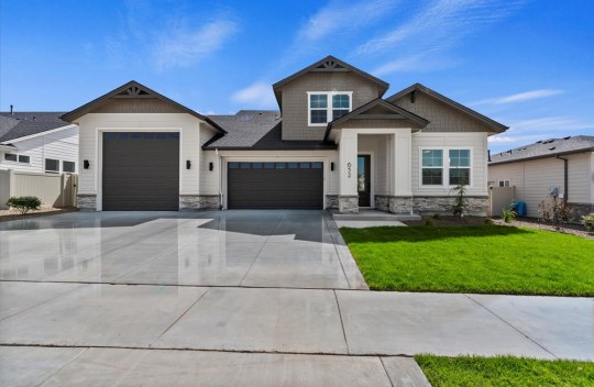 An exterior of a single story home with a gray and tan color scheme with dark accents of the roof and garage doors. There are stone details at the base of the home and green grass landscaping with rock pebbles and shrubs.