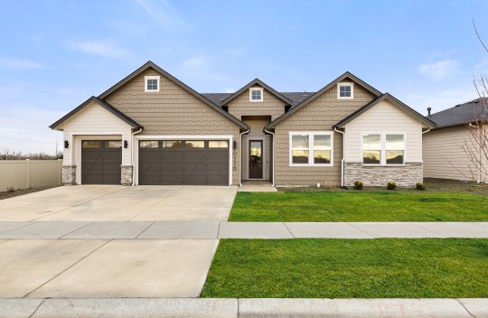 Exterior of a modern home with a tan, off- white and dark brown color scheme. Home has horizontal wood plank and wooden shingle architecture with 5 roof peaks, a 3 car garage, grass and tock landscaping.