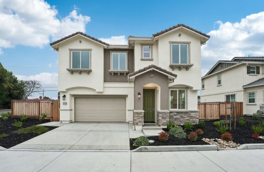 An exterior of a two story home with cream colored base paint with darker brown accents and stone detailing at the front entry. The yard is landscaped with dark mulch and small plants.
